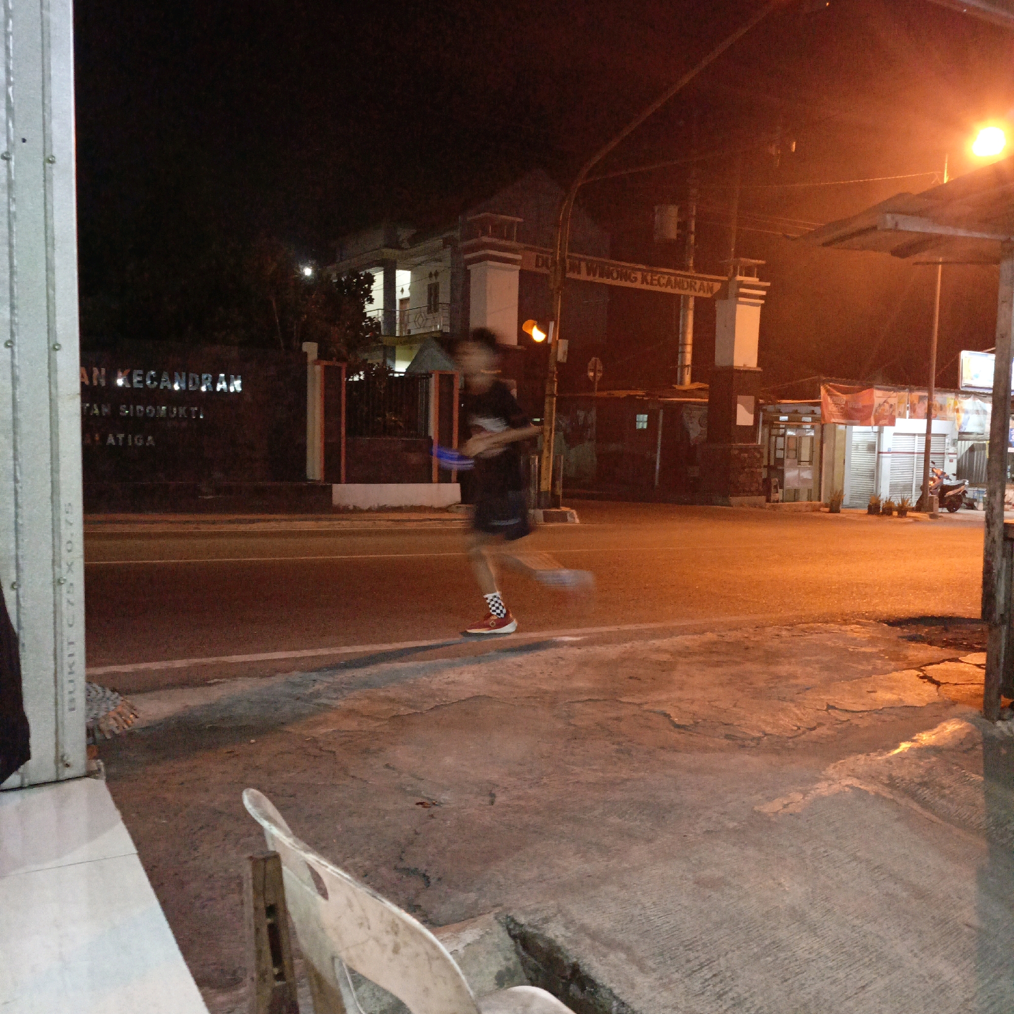 At night on a street in Salatiga, Indonesia, a blurry figure of a person is running. They are wearing a dark t-shirt, dark shorts, checkered socks, and red and white shoes. The person is holding a blue object in their left arm. A traffic light with a yellow light illuminated is visible behind them. In the background, there is a building with the text "DUSUN WIWONG KECANDRAN" illuminated. The street is paved and there is a sidewalk with some debris on it. A light pole with an illuminated light is on the right side of the image, casting a warm orange glow.