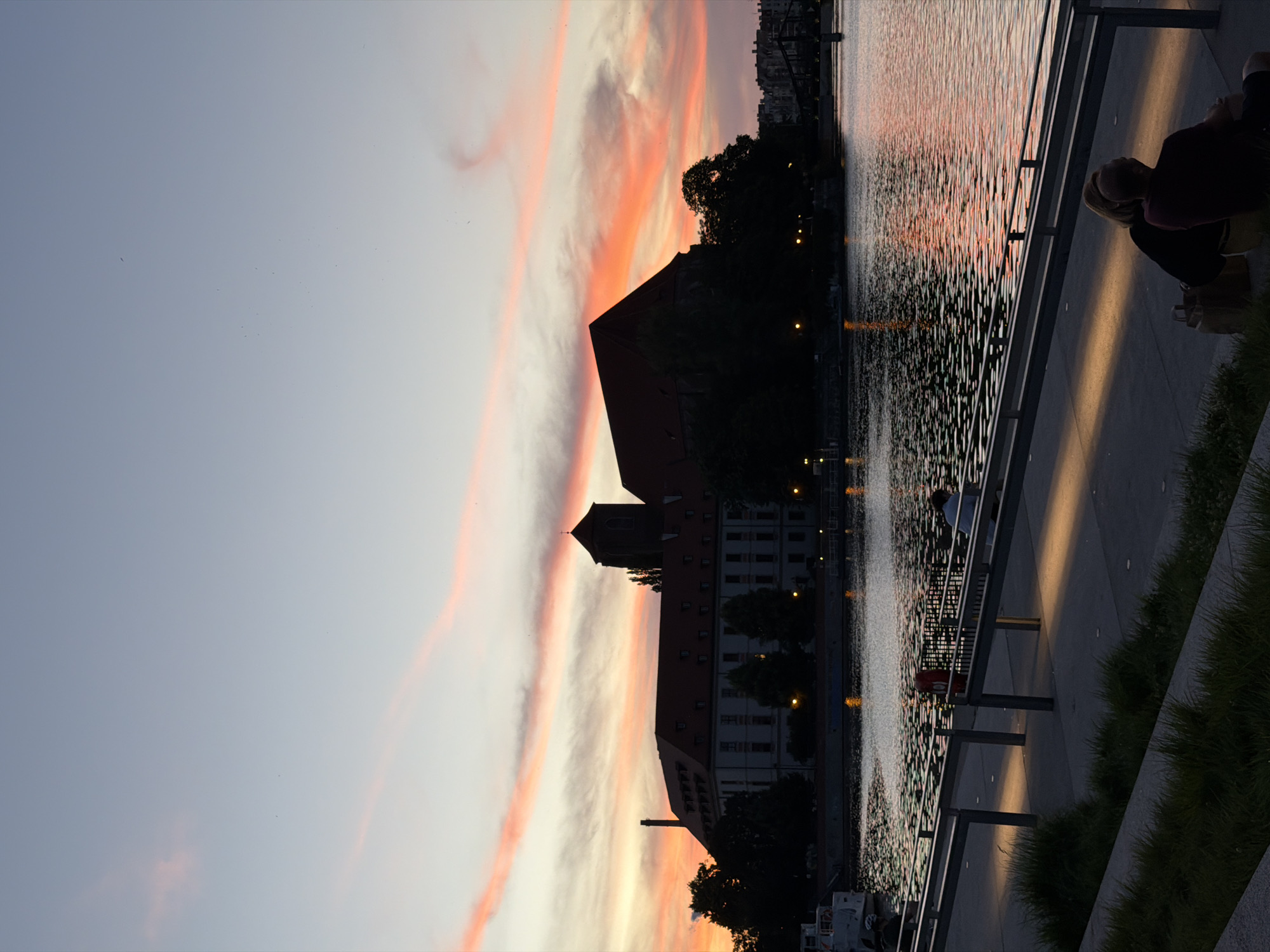 At dusk, a wide river reflects the colorful hues of a sunset, with streaks of pink and orange painting the sky. On the far bank, a large, imposing building with a red-tiled roof and a tall tower stands silhouetted against the sky. Trees partially obscure the building, and city lights twinkle in the distance. In the foreground, a paved walkway runs along the riverbank, bordered by metal railings. Several individuals are visible on the walkway, some sitting and others walking. The water's surface is rippled, and the light from the sunset casts a warm glow on the scene.