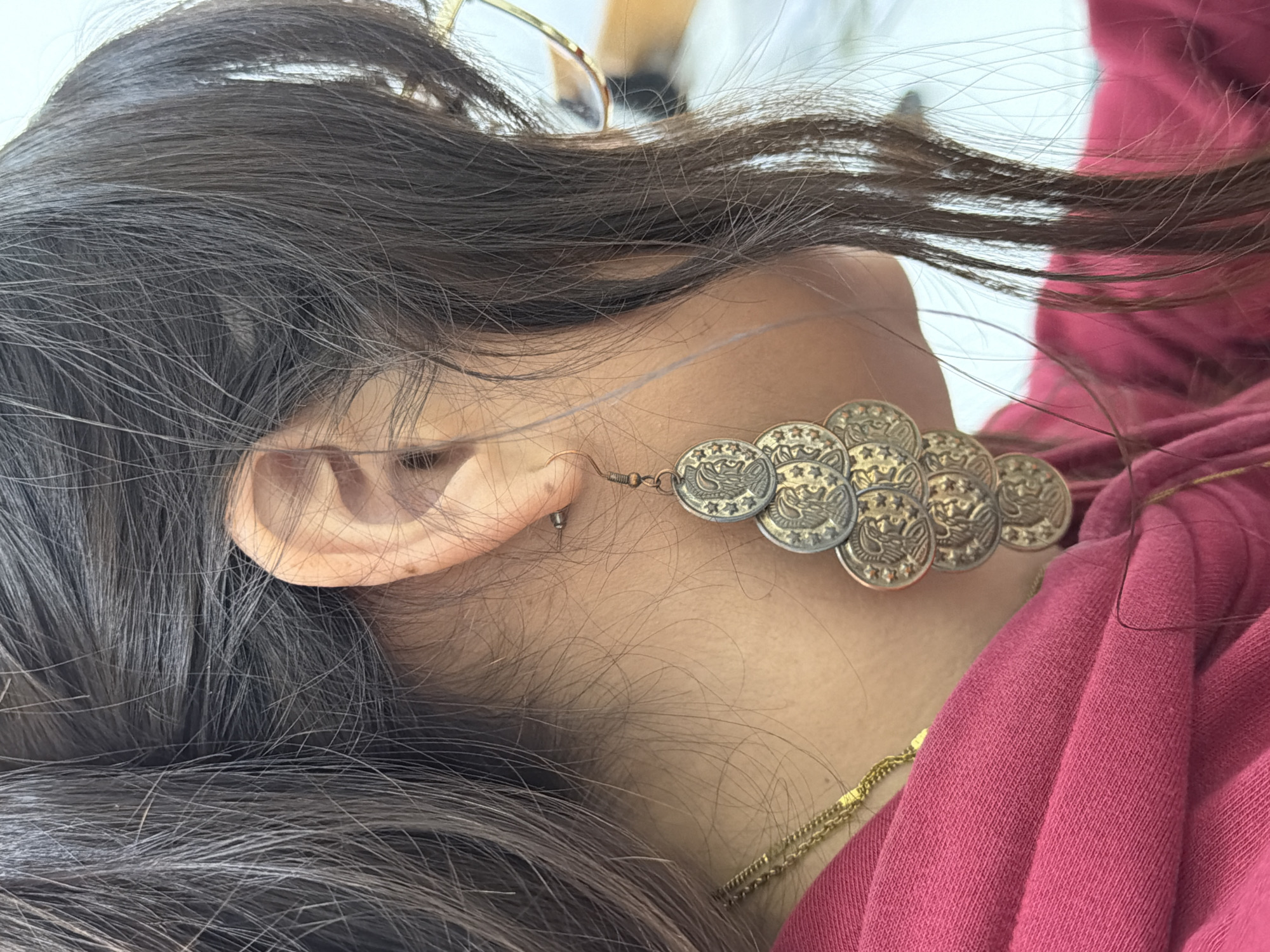 A close-up photograph shows a person's head and neck, presumably female. She has long, dark hair and is wearing a large, antique gold-colored earring made of multiple coin-like pieces. She also wears a delicate gold chain around her neck and a pink top. A pair of glasses is visible in her hair. The earring is the most prominent object. The setting of the image is San Andrés Cholula, Mexico.
