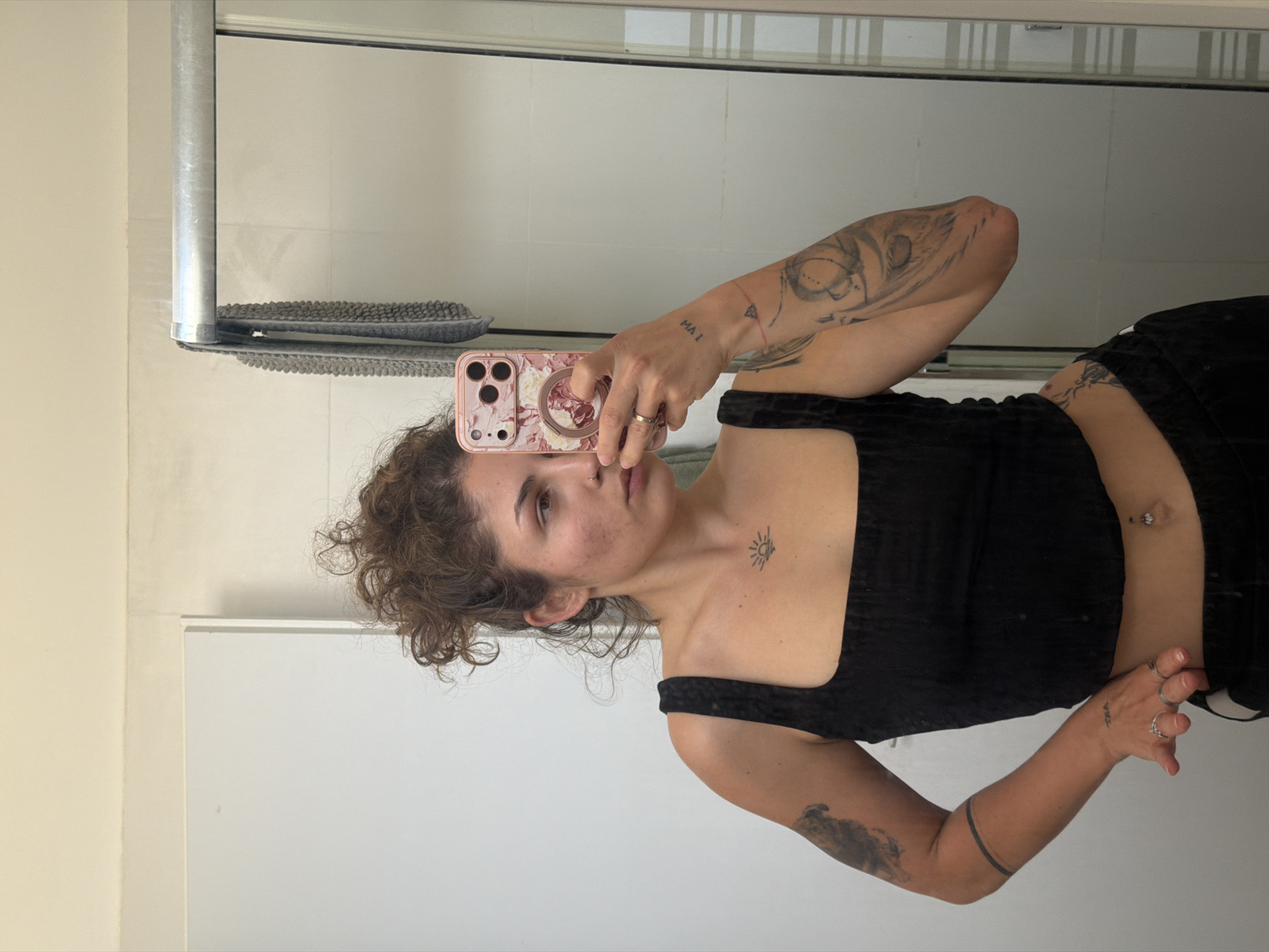 A woman is taking a selfie in a bathroom. She is holding a pink floral patterned iPhone in her right hand. The bathroom has white and gray tiled walls, with a metal shower rod and cleaning tool visible. The woman has curly brown hair, light skin, and is wearing a black top. She has tattoos on both arms, including on her left wrist a tattoo that reads "MA I." Other details include the presence of rings on her fingers and a navel piercing. The location is Kiryat Bialik, Israel.