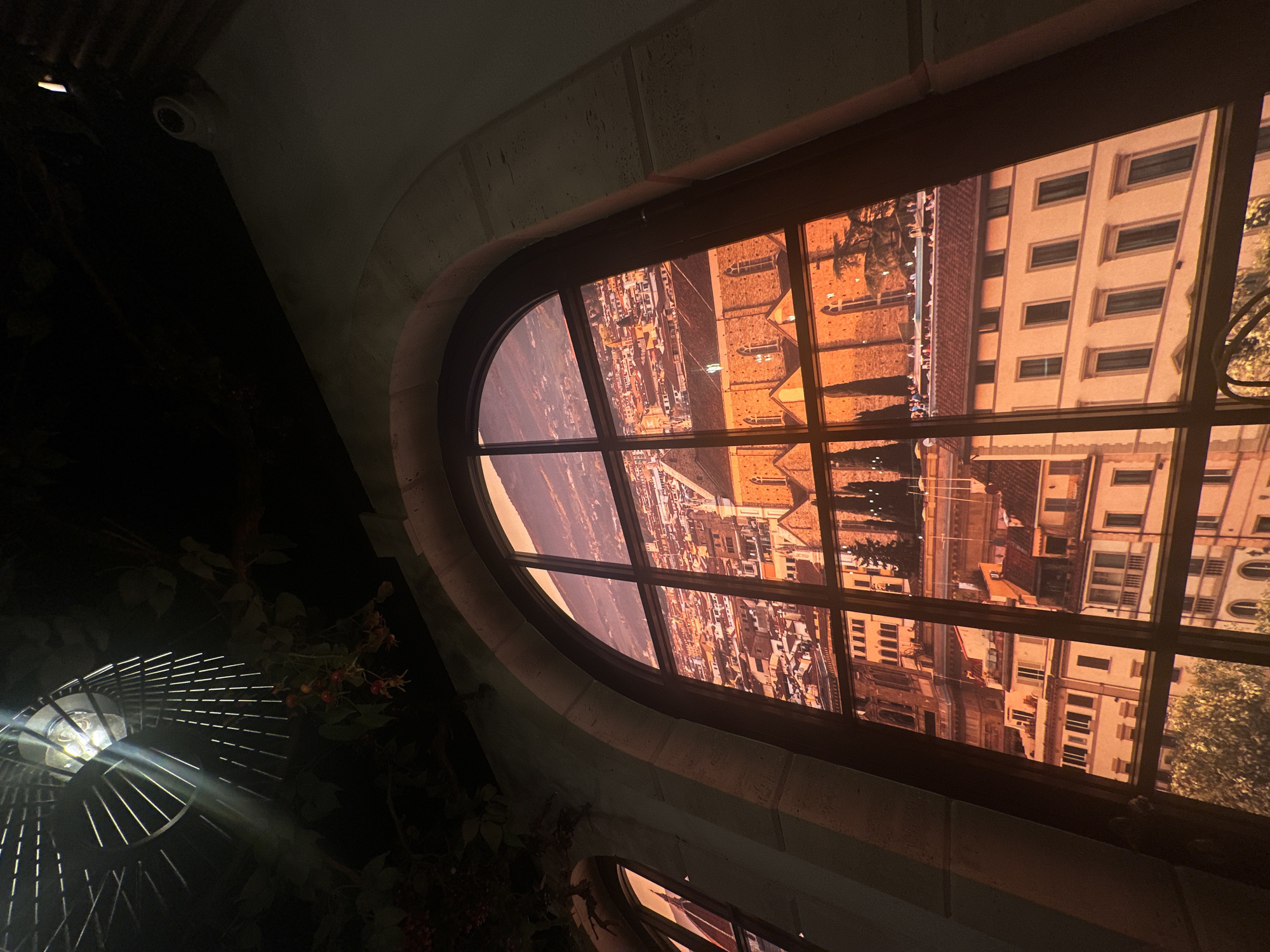 A low-angle shot of an arched window offering a vivid view of Florence, Italy. The architecture through the window is in warm tones, suggesting a sunrise or sunset. Outside the window, to the left, is a dark space with what appears to be plant life and a light fixture that resembles a caged fan. A security camera is visible on the upper left of the window frame. The frame of the window has a textured, stone-like appearance. The window itself is divided into several panes, creating a grid effect. A partial view of another window or archway is visible at the bottom.