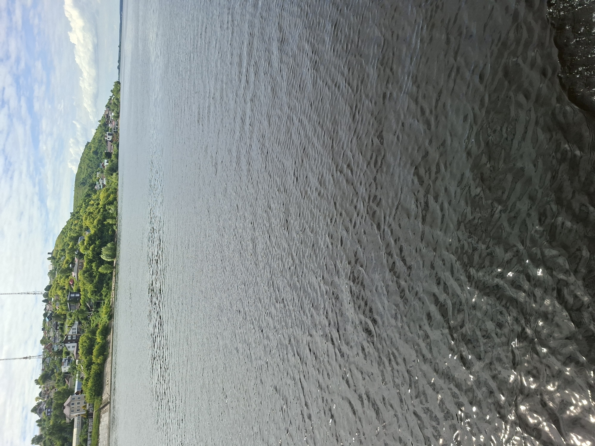 The image captures a wide expanse of a dark, rippling body of water, likely a river, under a partly cloudy sky. In the background, a verdant, hilly landscape is visible, dotted with numerous houses and buildings that form a settlement along the shoreline. The buildings are varied in size and style, with red, brown, and white roofs prominent amongst the greenery. Two tall radio towers are also visible on the highest point of the hill. The shoreline itself appears to be reinforced in some sections with concrete structures. The overall impression is of a tranquil, residential area situated along a significant waterway. The location is identified as Verkhny Uslon, Russia.