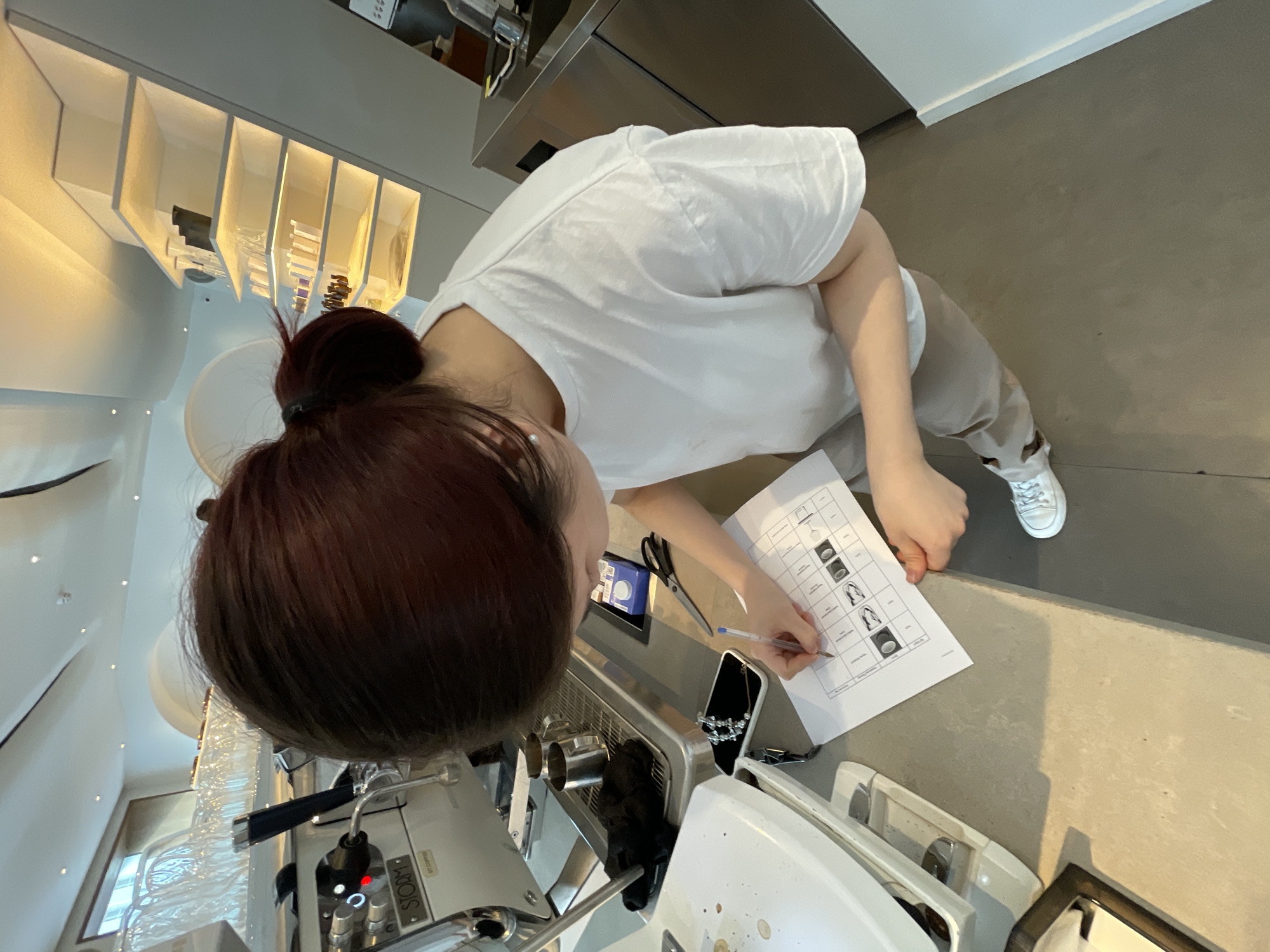 A person is writing on a paper with a pen, looking down at the document which appears to be a chart with pictures of different coffee cups. The person is wearing a white t-shirt and light-colored pants, with white sneakers. The scene is indoors, likely a cafe or coffee shop, with a professional-looking espresso machine visible on the left. Shelving units with various items, possibly coffee-related products or decor, are visible in the background. The overall impression is that of a barista or coffee enthusiast studying or making notes. The location is not specified in the image.