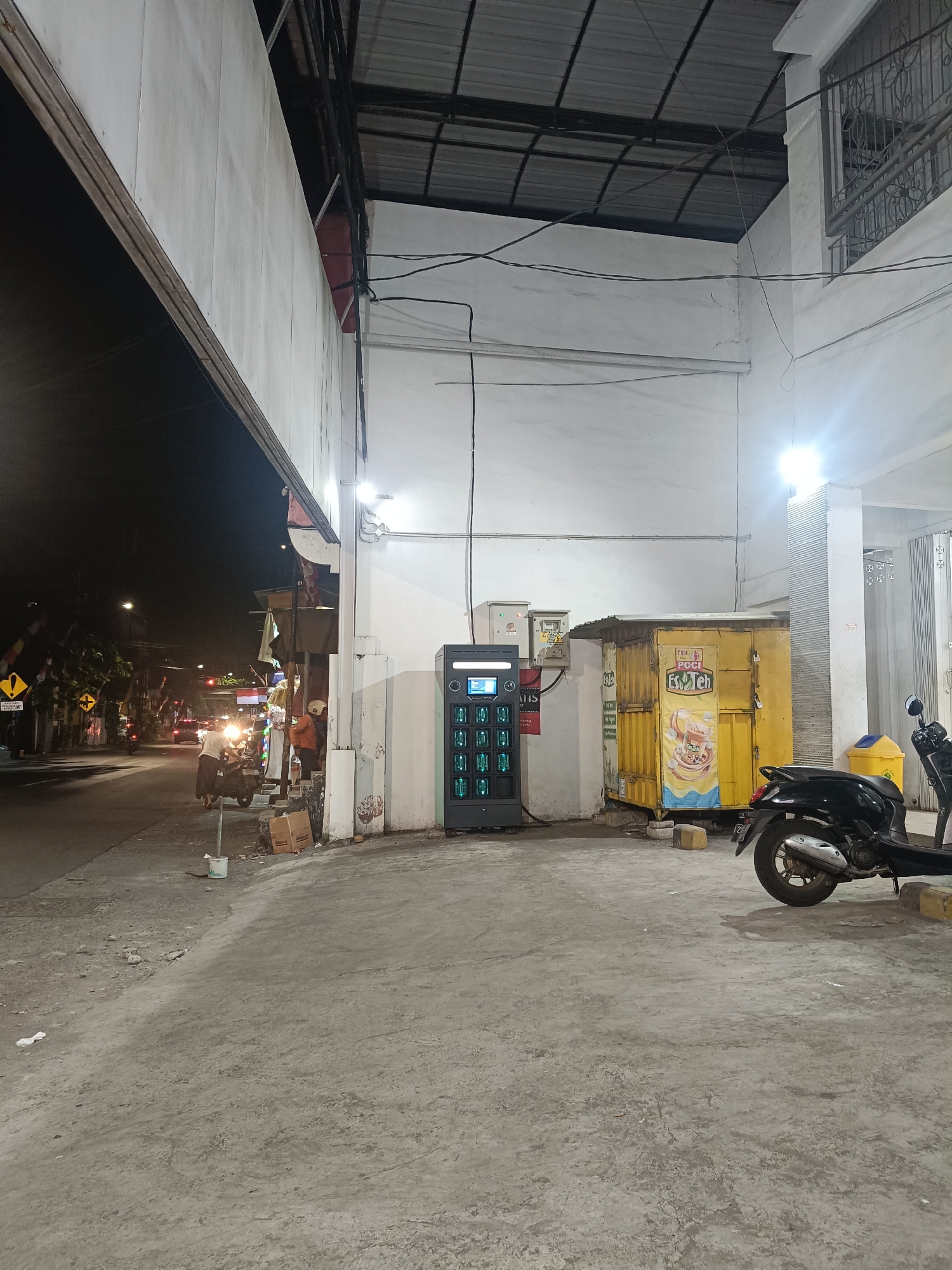 The image depicts a street scene at night in Surabaya, Indonesia, likely on a commercial street with moderate traffic. In the foreground, a concrete walkway leads to a white building. To the right of the walkway, a black scooter is parked. Adjacent to the scooter is a yellow kiosk with signage. In front of the kiosk is a tall, black vending machine with green illuminated compartments. Behind the kiosk, a white wall of a building is visible, with electrical conduits and lighting fixtures.
To the left of the walkway, the street is visible with passing vehicles and pedestrians. A yellow traffic sign with a warning symbol is present on the left side of the street. The street is illuminated by streetlights and the headlights of vehicles. The overall atmosphere suggests a typical urban evening in Surabaya, with a mix of commercial activity and transportation.