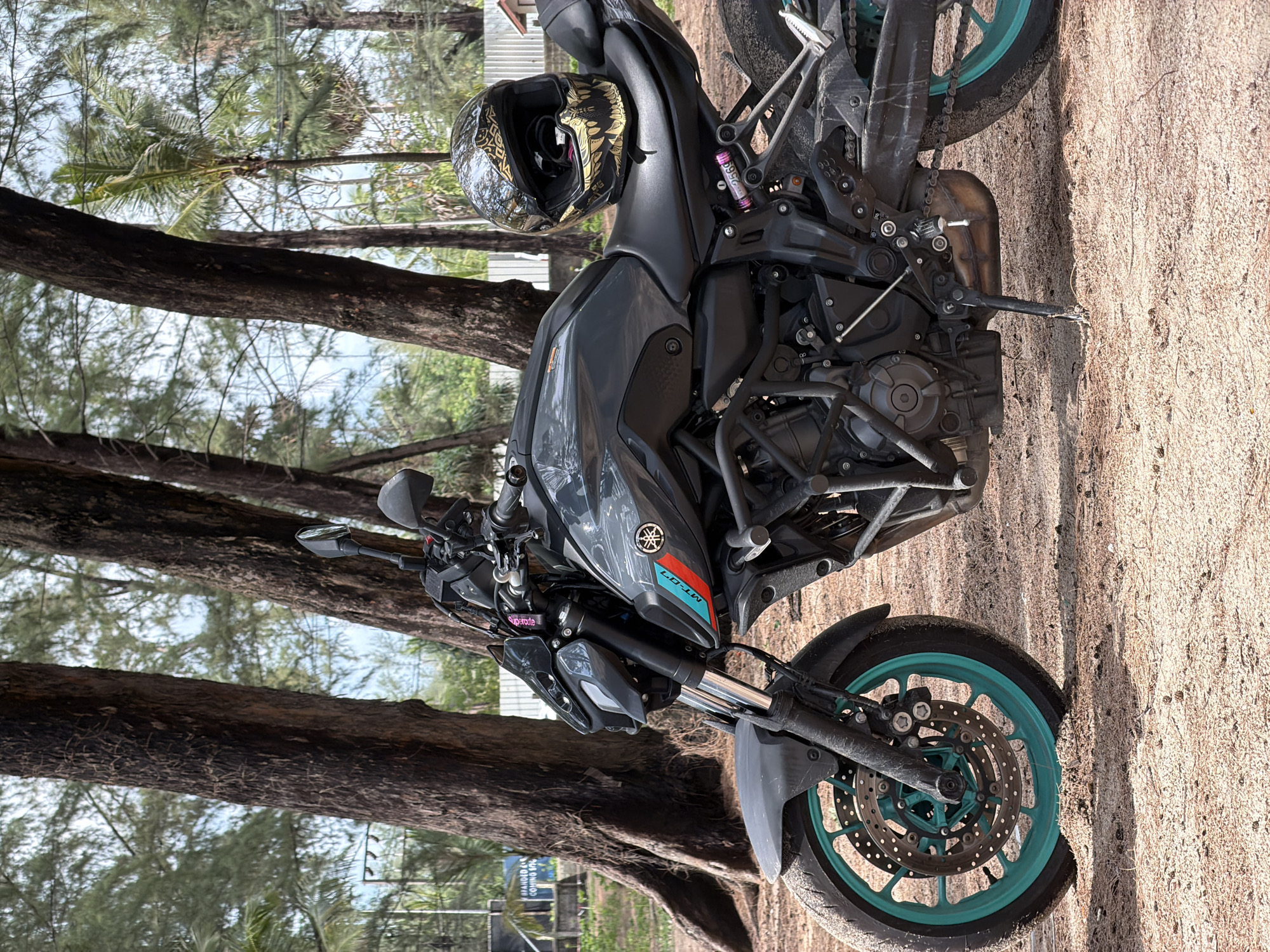 The image depicts a gray Yamaha MT-07 motorcycle parked on a sandy area with trees in the background. A black helmet with gold designs rests on the seat. The wheels of the motorcycle are turquoise. The location is Choeng Thale, Thailand, suggested by the presence of palm trees and the sandy ground resembling a beach. There are no people visible in the image.