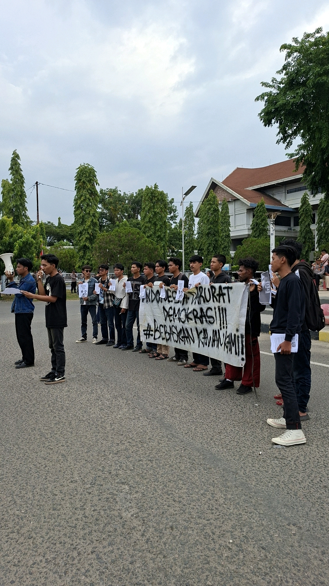 In Lhokseumawe, Indonesia, a group of young men are participating in a demonstration on an asphalt road. They are standing in a line, holding a large white banner that reads "INDONESIA DARURAT DEMOKRASI!!! #BEBASKAN KAWAN KAMI" which translates to "INDONESIA EMERGENCY DEMOCRACY!!! #FREE OUR FRIENDS". Some individuals are holding signs or papers. One person in the foreground on the left is holding a megaphone and appears to be speaking, while another individual next to them is also speaking into a microphone. The group is facing the camera, and the background shows a building with a reddish-brown roof, trees, and a cloudy sky. The overall atmosphere suggests a protest or public gathering.
