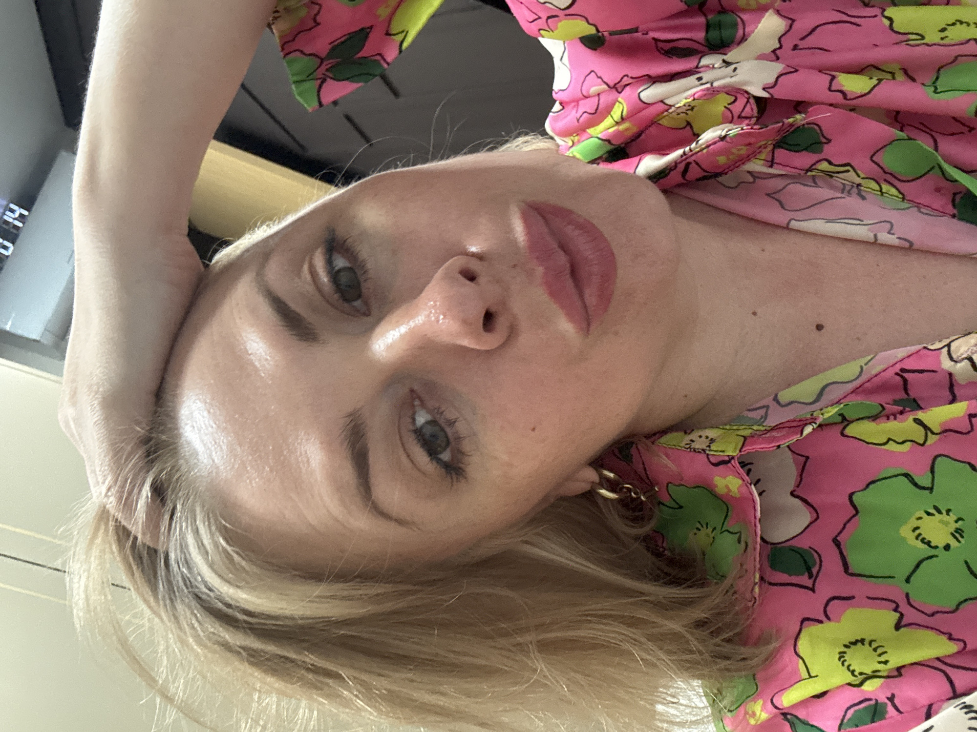 This is a close-up selfie of a blonde woman, possibly in her 30s or 40s, wearing a vibrant pink top with a floral pattern of green and white flowers. She has her left hand on her head, pulling at her hair, and her gaze is directed towards the camera with her lips pursed. Her makeup includes pink lipstick, mascara, and what appears to be blush or bronzer. She is wearing a gold hoop earring. The background is blurred, but a digital clock displaying "0 14" is visible in the upper right corner, suggesting an indoor setting. There are no discernible elements in the image that would indicate a specific location within the city of Sochi, Russia.