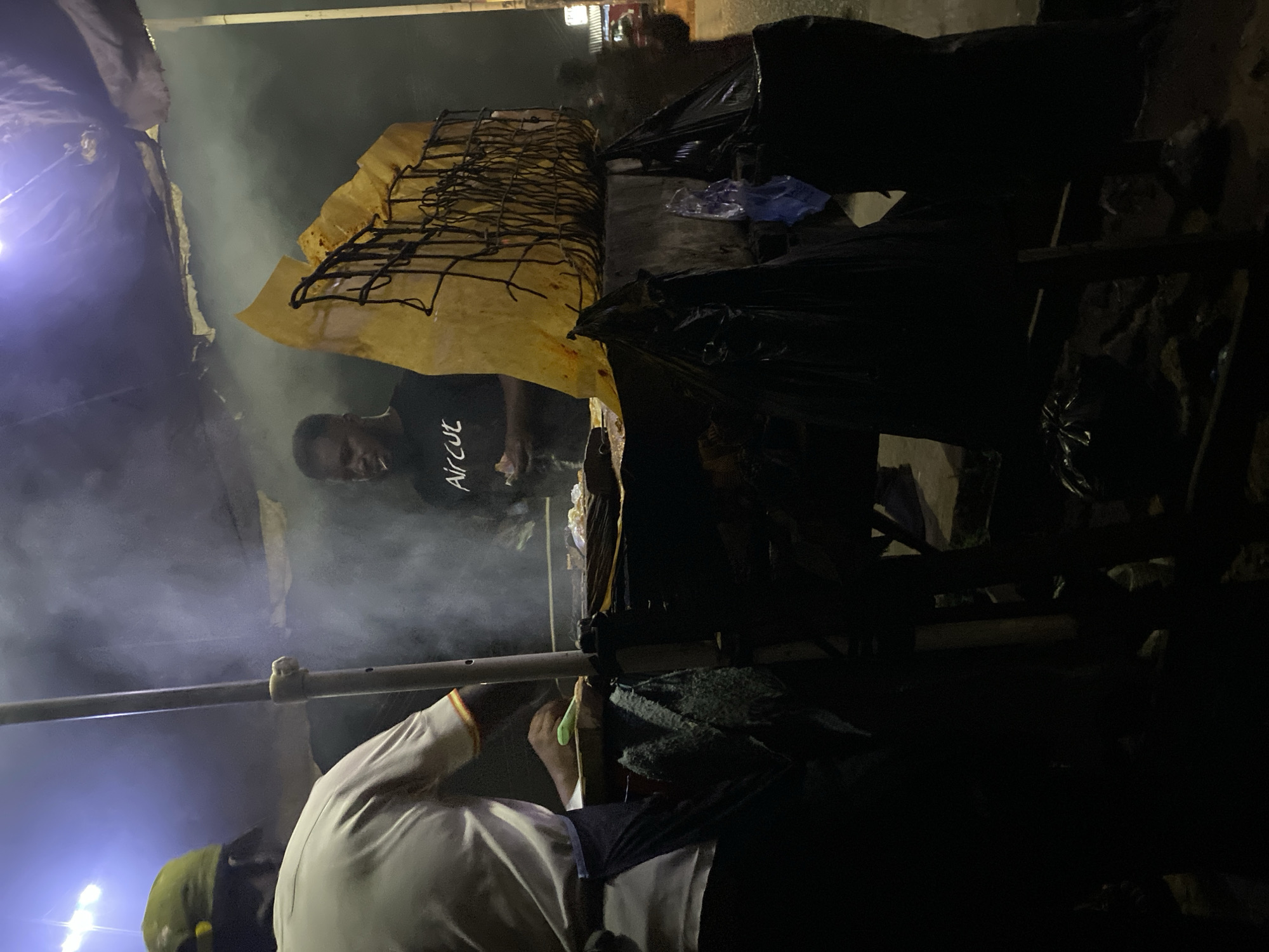 The image captures a vibrant, dimly lit street food scene at night in Bukuru, Nigeria. Heavy smoke billows upwards, suggesting active grilling or cooking over open flames.
In the mid-ground, a man wearing a black t-shirt with "Aircut" printed in white is visible behind what appears to be a food stall or counter. He stands to the left of a large metal grill rack, which is partially covered by a yellowish plastic or canvas sheet. He is looking towards the right, seemingly tending to the food being prepared.
In the immediate foreground, to the right, another individual is partially visible, wearing a light-colored shirt and a green head covering. This person is leaning over a surface, engaged in some preparation activity, obscured by the low light and angle. Further into the dark background on the right, another person is faintly discernible.
The setting is characterized by its rustic, outdoor nature. Several large black plastic bags are placed around the stall, possibly containing supplies or waste. A horizontal metal bar, likely part of the stall's structure, runs across the center of the frame. A bright, artificial light source illuminates the scene from the lower left, casting a glow on the dense smoke and some of the stall's elements, highlighting the bustling atmosphere of a night market. The overall impression is one of active food preparation late in the evening.