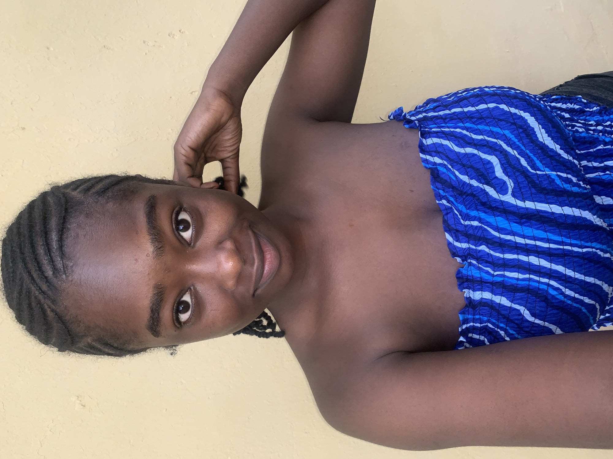 A young Black female is depicted in a head-and-shoulders portrait against a textured, light yellow wall, located in the city of Rafin Par, Nigeria. She gazes directly at the viewer with dark brown eyes and a subtle, closed-lip smile. Her hair is styled in dark, intricate cornrows. She is wearing a strapless, smocked top patterned with varying shades of blue and white wavy stripes. Her right hand, featuring dark nail polish, is raised and rests near the right side of her ear and neck.