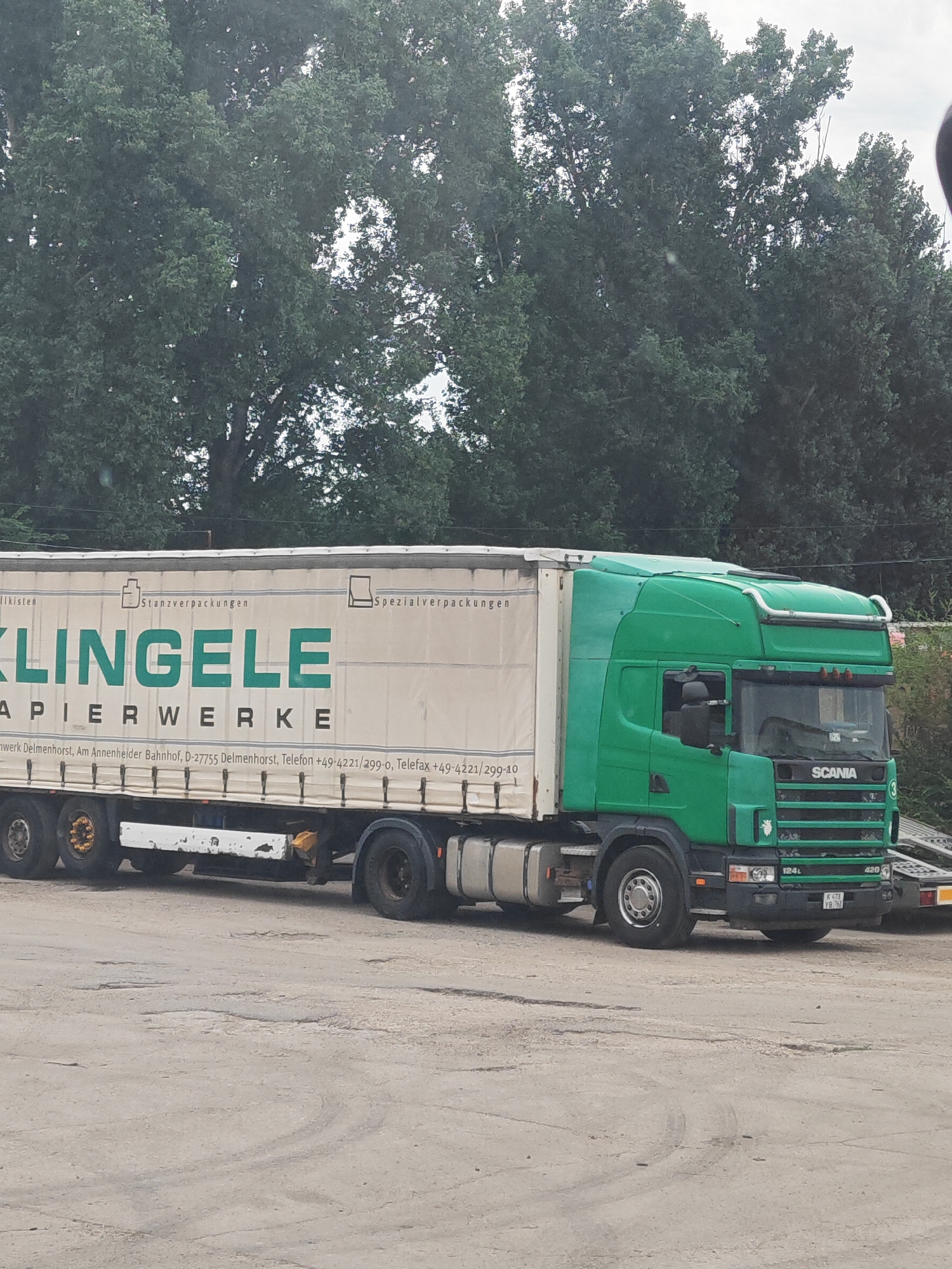 A green Scania truck with a white trailer is parked on a paved surface, likely a lot or industrial area, in Tolyatti, Russia. The trailer is branded with "KLINGELE PAPIERWERKE" in large green letters, indicating it is a paper factory. Text below the company name provides the address as "Delmenhorst, Am Annenheimer Bahnhof, D-27755 Delmenhorst" and contact information, including phone and fax numbers, suggesting this is a German company. The truck has its headlights and parking lights illuminated. Trees line the background of the scene, and the sky is overcast. The ground is cracked and uneven, with some tire marks visible.
