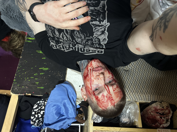 A person lies on their back, their face covered in fake blood. They are wearing a black t-shirt with a graphic design and a fitness tracker on their wrist. Below them, there's a wooden structure with clothing and a gruesome mask, possibly for a horror costume or prop. A section of a bed or upholstered surface is visible in the background. The overall impression is that of a staged or theatrical scene, possibly related to special effects or Halloween.