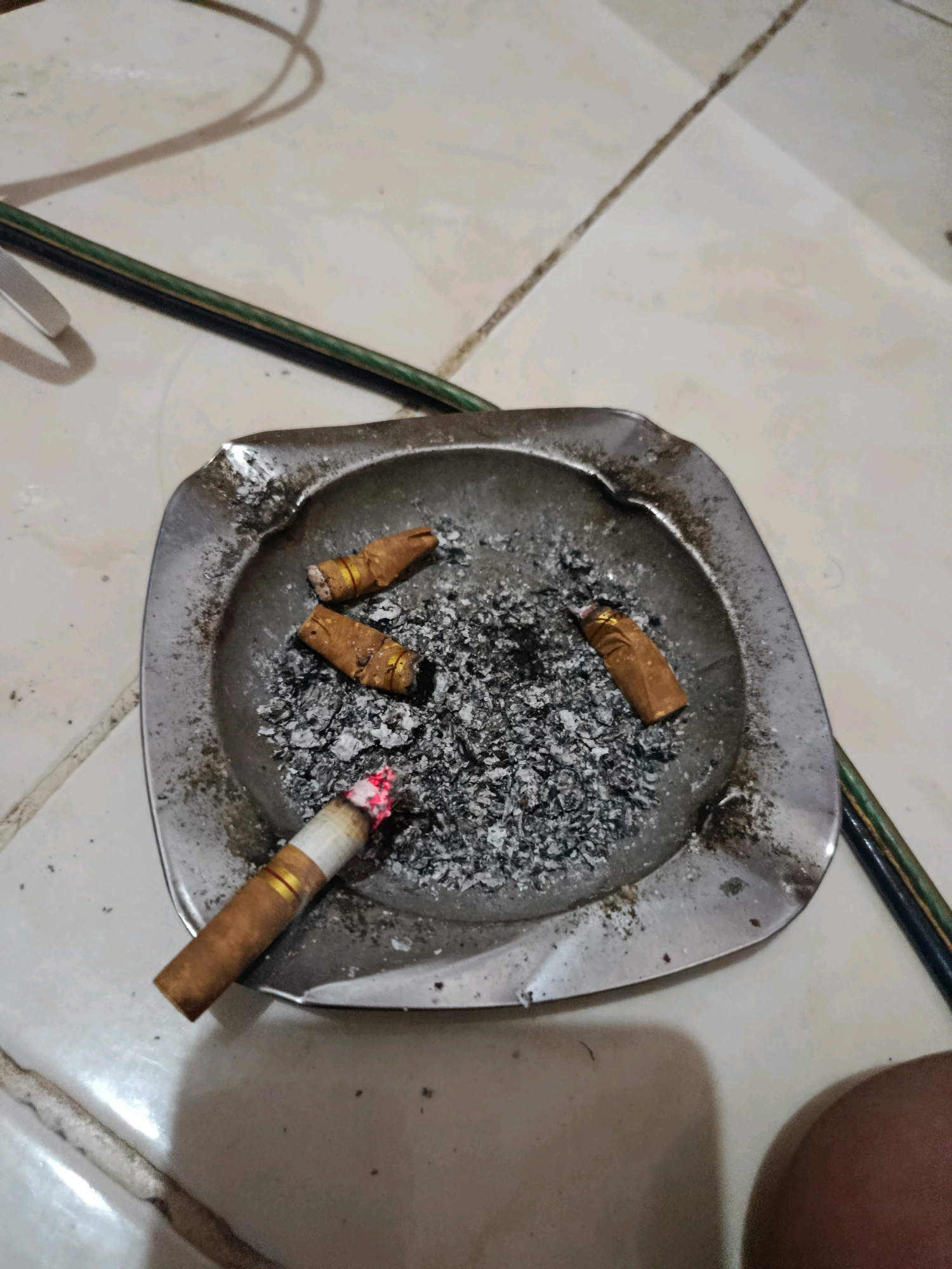 A close-up, overhead shot shows an ashtray on a tiled floor. The ashtray contains several cigarette butts and ash. One cigarette is still lit, with glowing embers. A green and black cable runs diagonally across the image behind the ashtray. The floor tiles are light in color and have dark grout lines. The scene appears to be indoors, likely in a residential setting.
