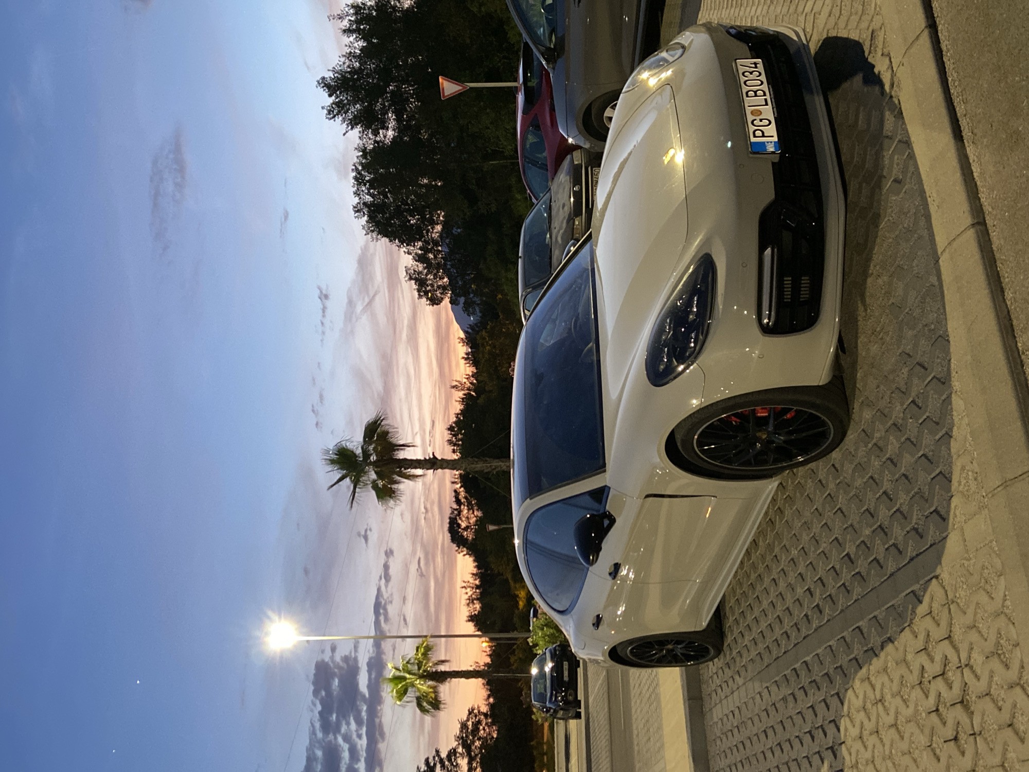 The image depicts a parking lot scene at dusk, located in Montenegro, as indicated by the "MNE" country code on the license plate. The license plate, reading "PG LB034", signifies registration in Podgorica, Montenegro.
In the foreground, a light grey Porsche Panamera (likely a 971 generation) is prominently parked on interlocking concrete pavers. The vehicle features black multi-spoke wheels, red brake calipers visible through the spokes, black exterior mirrors, and black trim around the windows and front grille/air intakes. Its headlights are off, but a reflective sheen is visible on the bodywork.
To the left and slightly behind the Panamera, a dark-colored Chevrolet Camaro is parked. Further in the background, other vehicles are visible, including a red hatchback and a dark sedan, though their precise makes and models are less discernible. A red triangular "Give Way" (or "Yield") sign is positioned on a pole to the right, behind the parked cars.
The environment includes two distinct palm trees, one to the left illuminated by a bright streetlamp, and another to the right. A dense line of trees forms the background, with the faint silhouette of distant mountains visible above them on the right side. The sky displays a sunset gradient, transitioning from warm orange, pink, and purple hues near the horizon to a clear blue overhead. Scattered clouds are present, some reflecting the sunset colors. Two faint white dots, possibly stars or planets, are visible in the upper left portion of the sky. The ground consists of patterned concrete pavers with a central drainage channel. No people are visible in the scene.
