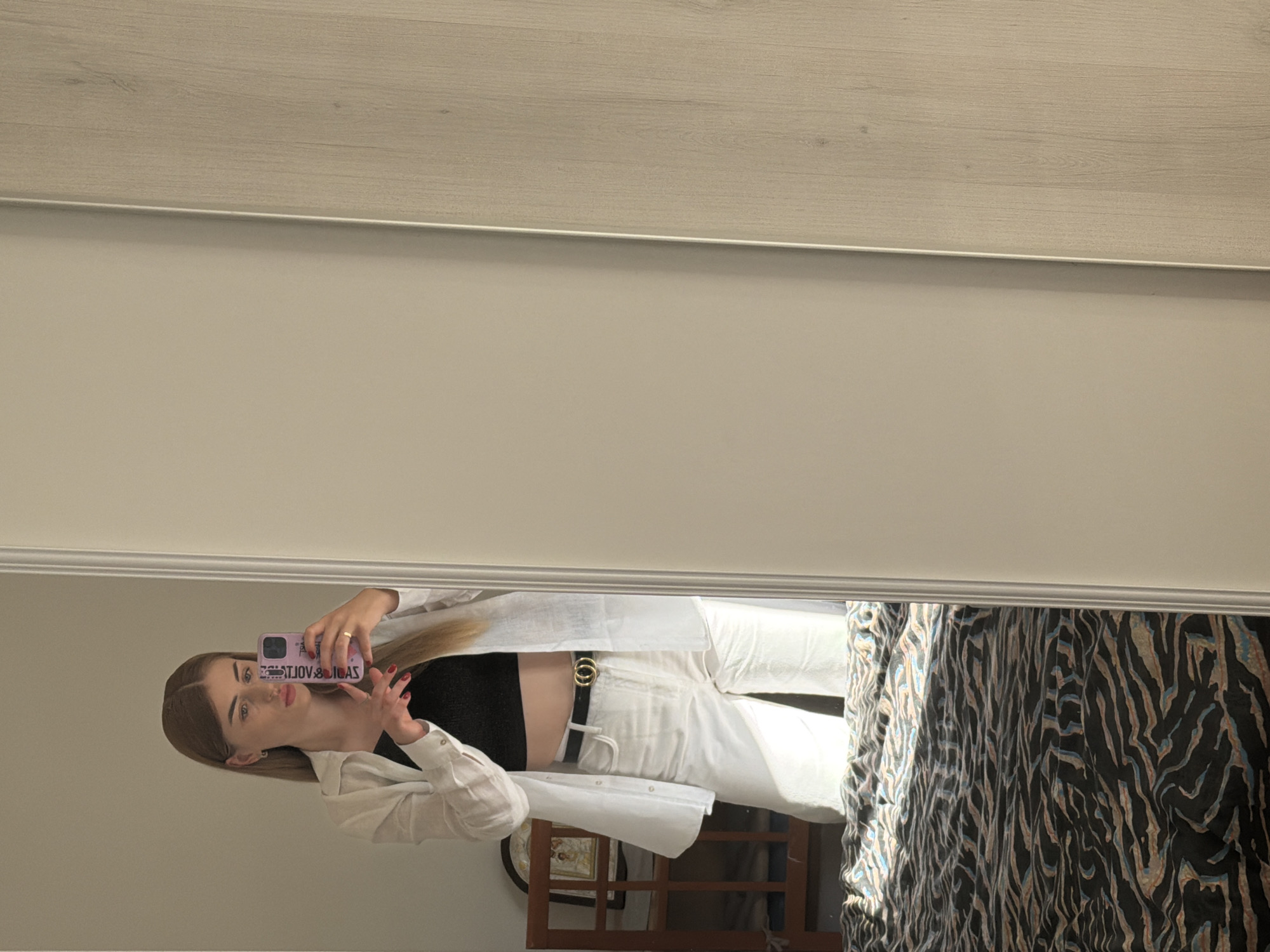 A young woman is reflected in a mirror, taking a selfie with her phone. She is wearing a black crop top, white pants, a white linen shirt, and a black belt with a gold double G buckle. Her hair is long and blonde, and she is wearing subtle makeup. The background of the reflection shows a wall with a light wood panel above a cream-colored section. To the right of the woman in the reflection, part of a bed with a dark comforter featuring a zebra-like pattern is visible. The woman's phone case is pink and has text that appears to read "VOLTAGE" and "SUVI". The lighting suggests it is daytime, and the scene appears to be indoors, likely in a bedroom. The woman's expression is neutral, and she is looking directly at the camera.