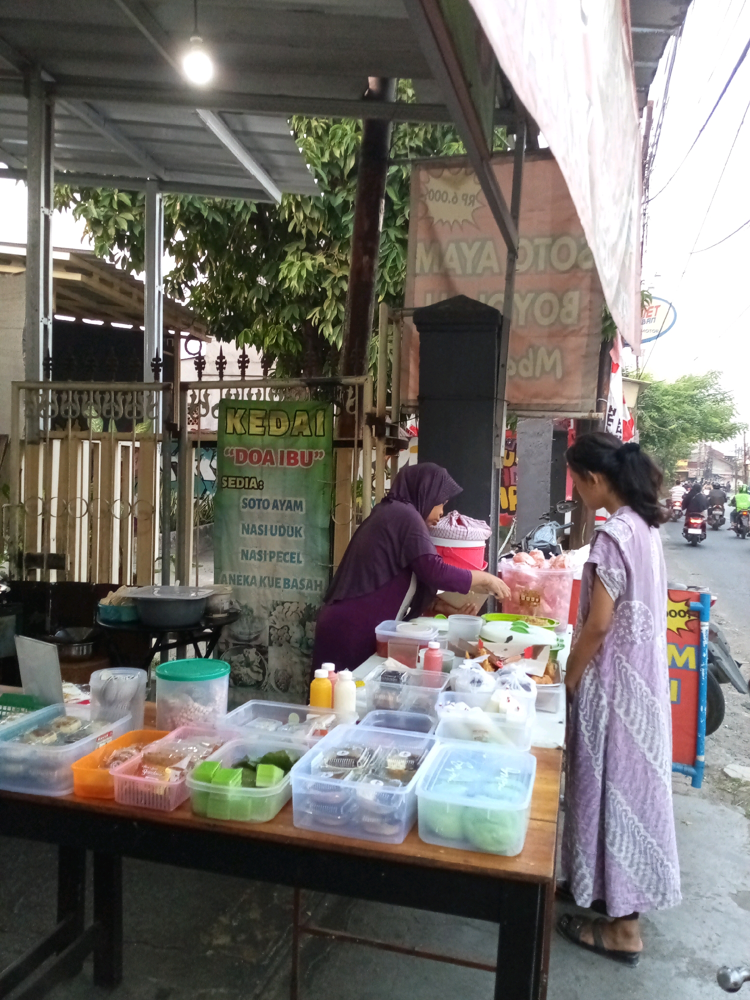 A street food vendor is serving a customer in front of a food stall named "Kedai DOA IBU" in Bekasi, Indonesia. The vendor, wearing a purple hijab and tunic, is standing behind a table laden with various food containers. She is in the process of handing over a plastic container of food to a customer, a young woman wearing a long, patterned purple dress and sandals. The food stall is situated on the side of a street with traffic, indicated by passing motorcycles in the background. A green sign behind the vendor lists menu items like "Soto Ayam," "Nasi Uduk," and "Nasi Pecel." The environment appears to be a typical urban setting with overhead awnings and buildings visible.