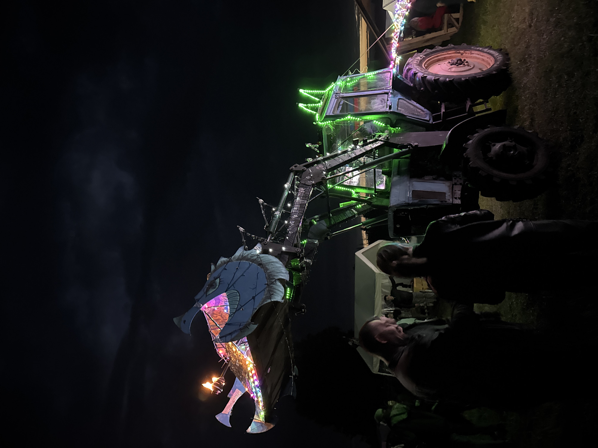 The image depicts a nighttime scene in Большая Кивара, Russia, featuring a modified tractor as the central illuminated spectacle. A dark-colored agricultural tractor, equipped with large tires, has its front loader arm extensively adapted to form the neck and head of a dragon. The dragon's head, constructed from metal panels and painted in muted tones, is adorned with multi-colored string lights (red, green, blue, yellow, pink) and features two open flames at its snout. The tractor's cabin and front structure are outlined with green LED lights, including illuminated "spikes" on the roof.
The event takes place outdoors on a grassy field under a dark, cloud-strewn night sky. Multiple individuals are present, observing the illuminated tractor. In the immediate foreground, two people are partially silhouetted: a man with a beard in a dark jacket, facing right, and another person with their back to the viewer, also in a dark jacket. To the far left, a person in a green outfit is partially visible. In the midground, near white tents, additional people are visible, mostly as silhouettes. Ambient lighting is provided by separate strings of multi-colored lights draped across the scene, particularly on the right side, where a person in red is seated on a visible wooden structure. The overall lighting is low, highlighting the artificial illumination of the central subject.