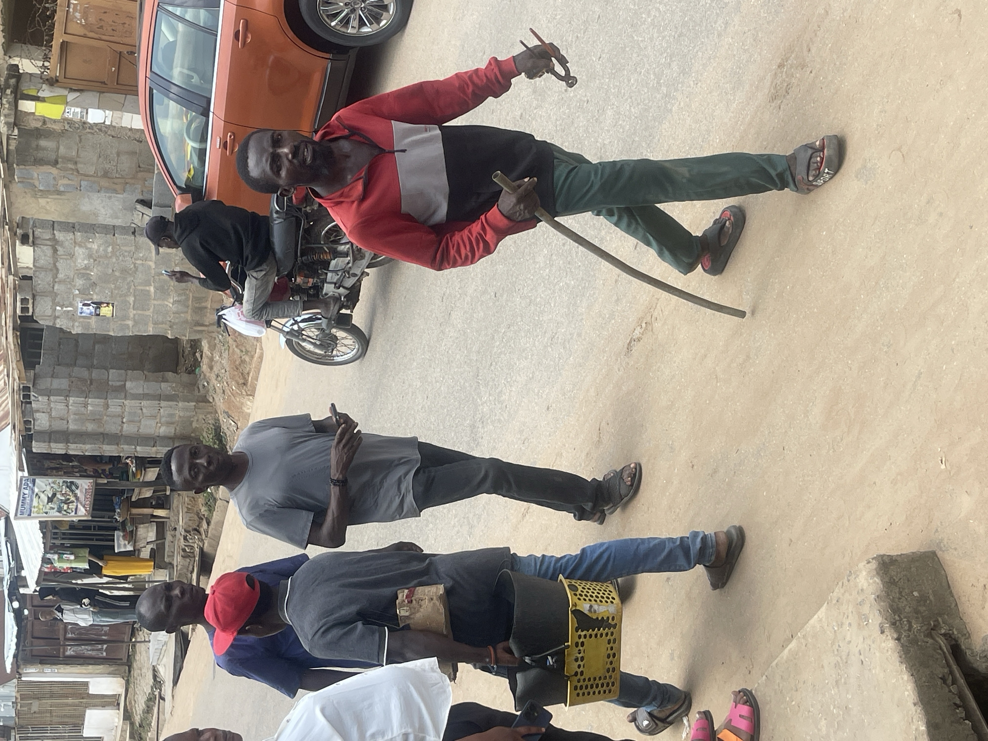 The image captures a street scene in Abuja, Nigeria, where several individuals are walking along a dusty road. In the foreground, a man in a red, grey, and black zippered jacket and green pants carries a long, thin pole in his left hand and a pair of pliers in his right. To his left, a person wearing a dark blue shirt and a red cap is walking, and another individual in a grey t-shirt and dark pants is to the far left. Further back, a person in a grey t-shirt and dark pants walks with a yellow basket containing dark items. In the background, an orange SUV is parked, and a motorcycle with a rider wearing a black hoodie and cap is visible. The environment appears to be an urban street with buildings and shop fronts lining the road, some with corrugated metal roofs.