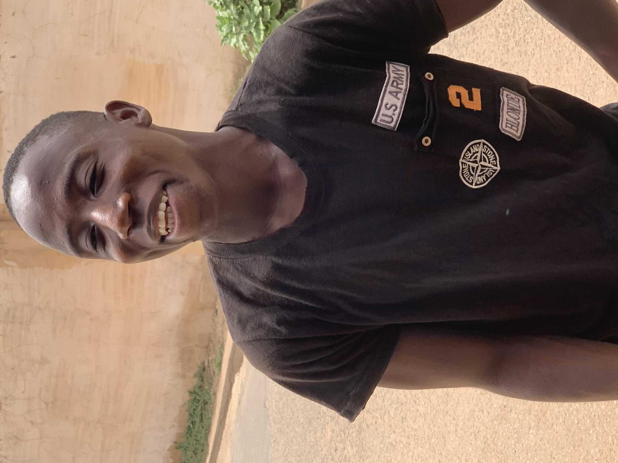 A young man is smiling at the camera while standing outdoors. He is wearing a black short-sleeved polo shirt with several patches on the upper chest. The patches include: "U.S. Army," the number "2," "Blonde," and a "Stone Island" logo. The background includes a light brown wall, green vegetation, and what appears to be a paved area. Given the presence of vegetation, the light brown wall and the paved area, it is likely a residential area. The exact location within Jos, Nigeria, cannot be definitively determined from the image, but it appears to be an outdoor, possibly residential, environment in daylight.