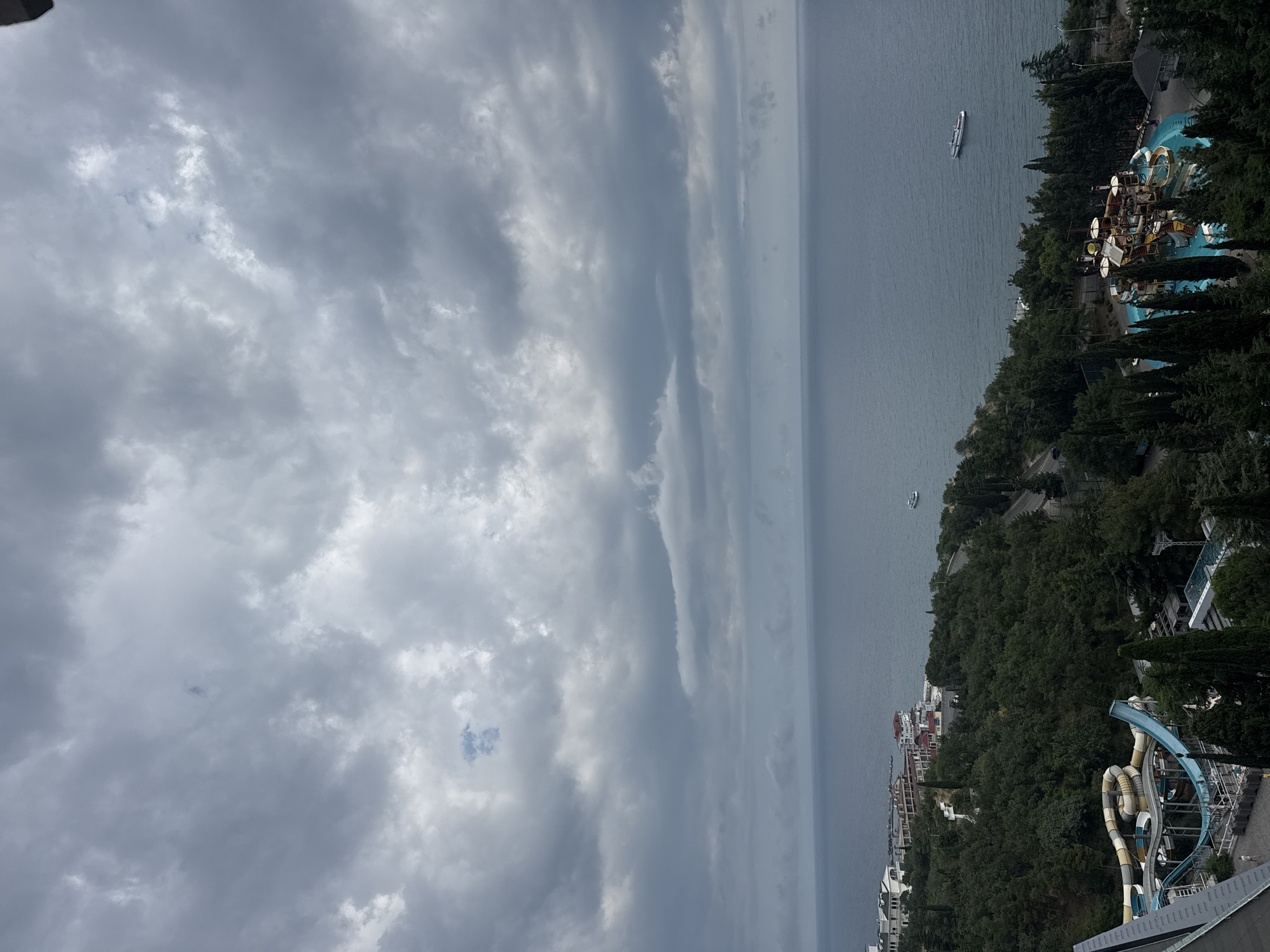 The image depicts an overcast sky over a coastal scene. In the foreground, a portion of a building is visible on the left. Below, a water park complex with blue and yellow slides is situated amidst dense green trees. To the right of the water park, another aquatic play area with multiple structures and a pool is visible, also surrounded by trees. Further inland, buildings and a road are nestled within the greenery. The sea stretches out to the horizon, with a white ship sailing on its surface. The location is identified as Yalta, Ukraine.