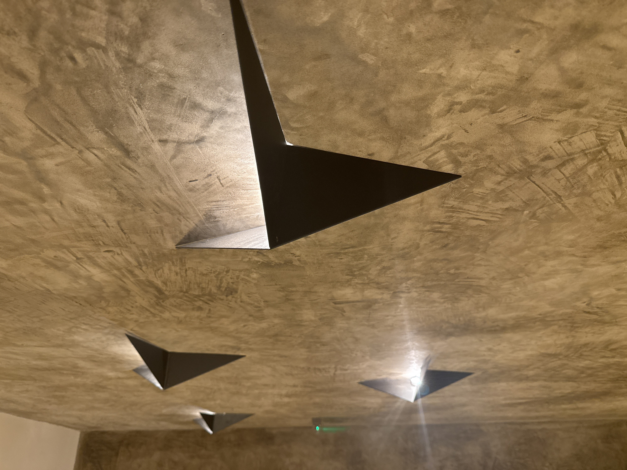 The image displays a textured concrete wall with four triangular wall sconces mounted on it. The sconces are made of a dark material, possibly metal, and are angled outwards, casting light onto the wall. The wall itself has a rough, mottled finish in shades of beige and gray. The lighting is dim, with the sconces being the primary light sources. A small, green indicator light is visible on the wall between the two leftmost sconces. The image is taken at an upward angle, suggesting the sconces are mounted at a moderate height on the wall. This scene is likely from the interior of a modern building or establishment, possibly a restaurant, hotel, or office space. Without further context, the specific location in Varna, Bulgaria cannot be determined from the image alone.