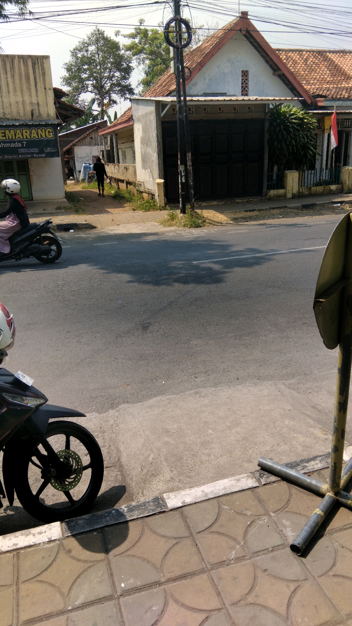 The image shows a street scene in Proyonangg, Indonesia. A person is riding a motorcycle in the foreground, with another motorcycle visible to the left. In the background, there is a building with a tiled roof and a large black gate. A man is walking on the sidewalk in front of the building. There are trees and power lines in the background, indicating an urban environment. The weather appears to be sunny.