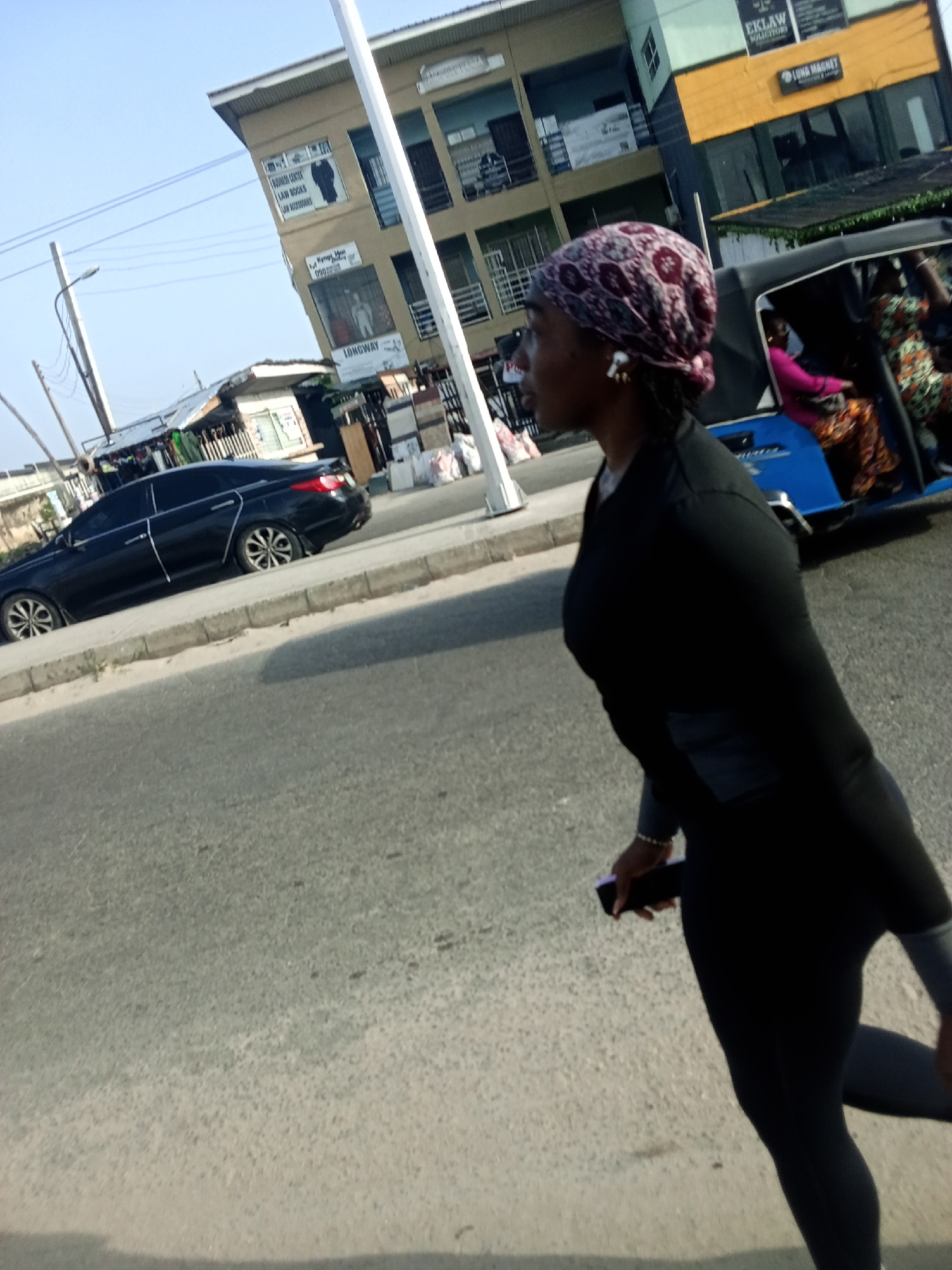 The image shows a street scene in Ado, Nigeria. A woman wearing a dark athletic outfit and a patterned headscarf is walking on the road, holding a mobile phone. Behind her, to the right, is a blue motorized tricycle (keke) with passengers inside. Further in the background to the left, a black car is parked along the curb. Buildings are visible on the opposite side of the street. These buildings appear to house businesses, as signage is visible. One sign reads "BUSINESS CENTER LAW BOOKS LAW ACCESSORIES" and "LONGWAY." There is also a sign stating "E KLAW SOLICITORS" and "LONA MAGNET". A utility pole is to the left of the buildings. The street is paved, and it appears to be daytime.