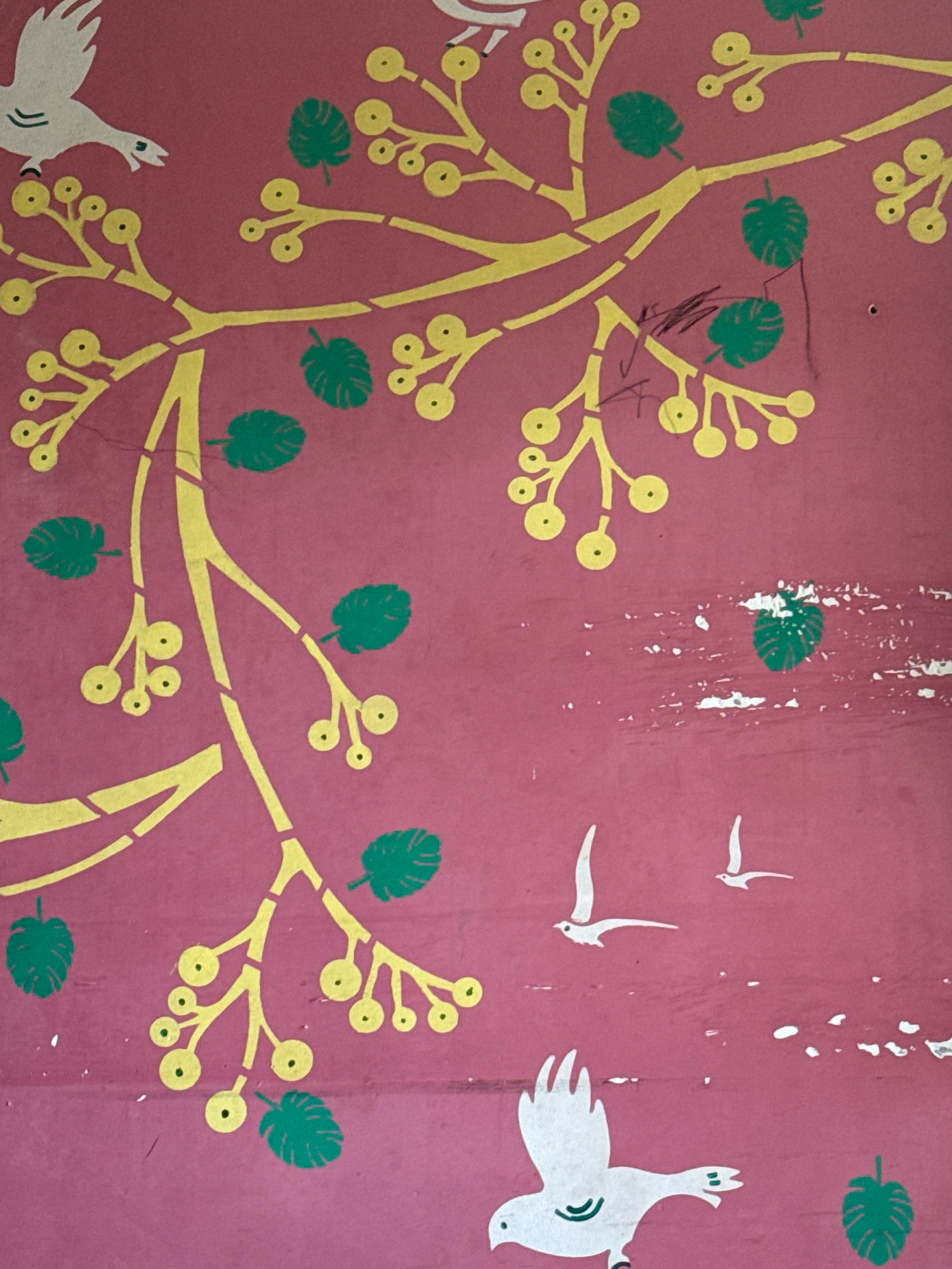 The image shows a close-up of a wall with a stylized illustration of a tree with yellow branches and green leaves and yellow berries. Two white birds are depicted in flight, one larger than the other. A third white bird, with green markings on its body, is positioned near the bottom of the frame. The wall is a reddish-pink color and shows signs of wear and tear, with some white scuff marks and a small hole visible. There are also some dark scribbles in the upper right portion of the image, possibly from a pen or crayon. The image is a photograph of a mural or painted design on a wall, located in Dubacherla, India.