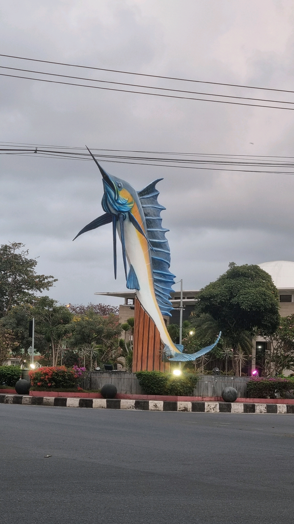 The image shows a large, colorful sculpture of a marlin in the center of what appears to be a roundabout or traffic island in Desa Pagandaran, Indonesia. The sculpture is depicted leaping out of the water, with its dorsal fin extended and its bill pointed upwards. The marlin is predominantly blue and white, with accents of orange and yellow. It is mounted on a wooden pedestal.
The roundabout is surrounded by lush greenery, including trees and flowering bushes. There are also several round, dark-colored decorative balls placed around the base of the sculpture. In the background, a building with a domed roof is visible, along with some trees and more vegetation. The sky is overcast, suggesting a cloudy day. Power lines are strung across the top of the image. The road surface in the foreground is asphalt, with a black and white striped curb marking the edge of the roundabout. There are no people visible in the image.