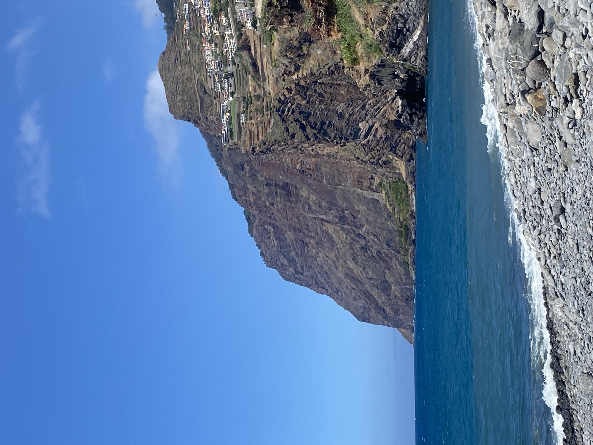 The image depicts a coastal landscape with a prominent cliff face overlooking a rocky beach and the Atlantic Ocean. The cliff, a sheer, textured rock formation, rises dramatically from the water, its surface marked with various shades of brown and grey. Clinging to the upper slopes of the cliff are terraced agricultural fields and a cluster of white buildings, indicative of a settlement. These buildings, with their red-tiled roofs, are spread out along the contours of the land, suggesting a village or town perched on the hillside.
The ocean is a vibrant, deep blue, with gentle waves breaking against the shore. The beach itself is composed of smooth, rounded grey and white pebbles. The sky is a clear, bright blue, with a few wispy white clouds present. The scene appears to be captured on a sunny day, with strong natural light illuminating the landscape. The context of Câmara de Lobos, Portugal, suggests this is likely a view of the dramatic coastline characteristic of Madeira. There are no people or animals visible in the image.