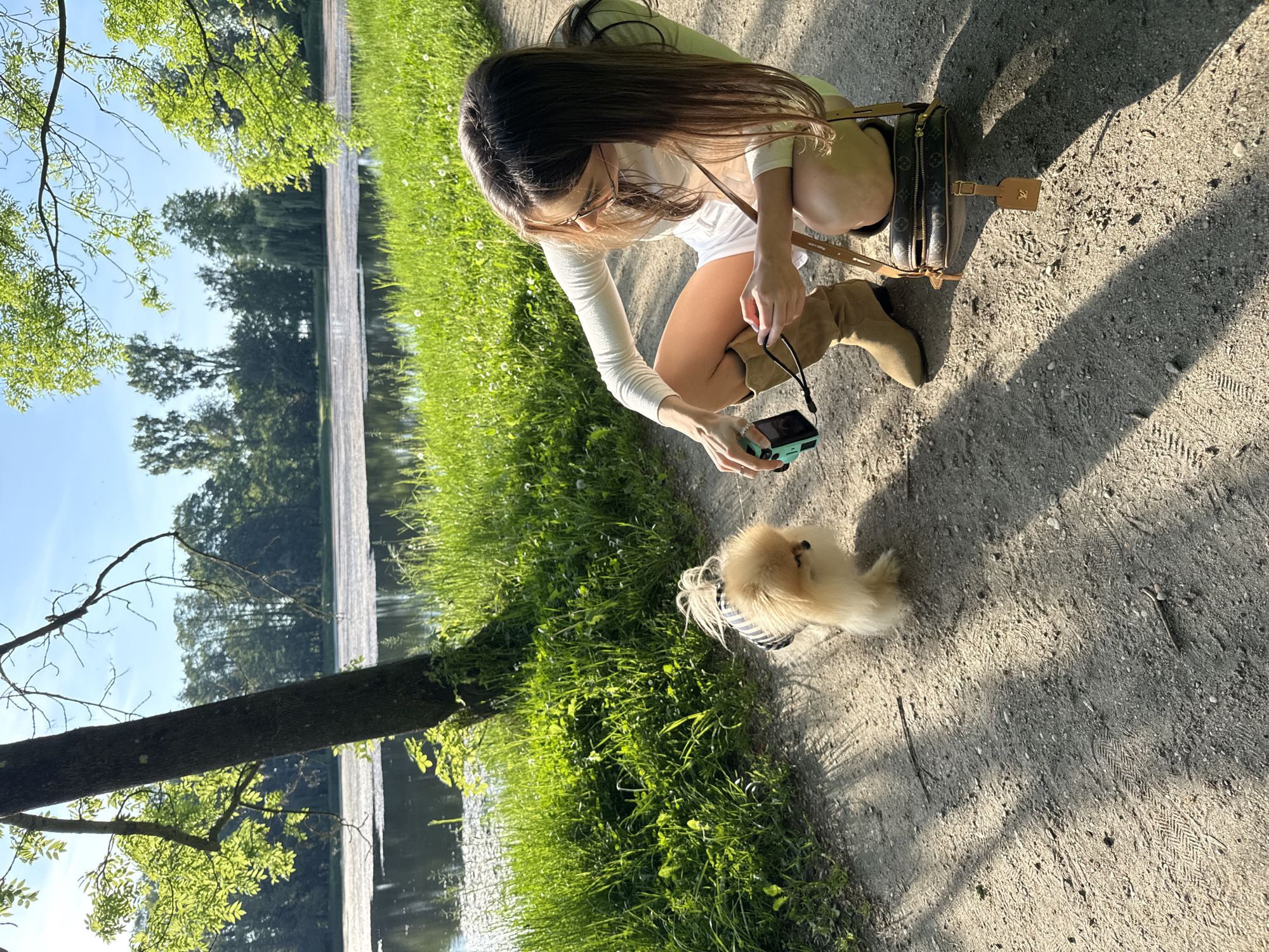 A woman is kneeling on a dirt path next to a small dog, holding a camera and taking a picture. The woman is wearing a long-sleeved white shirt, white shorts, and tan boots. She has long brown hair. A Louis Vuitton handbag sits behind her. The dog is a small, tan Pomeranian wearing a striped shirt. To the left, there is a body of water with a tree trunk overhanging the water. The background is a grassy area with trees. The scene is outdoors and well-lit. The location is Unknown, Unknown.