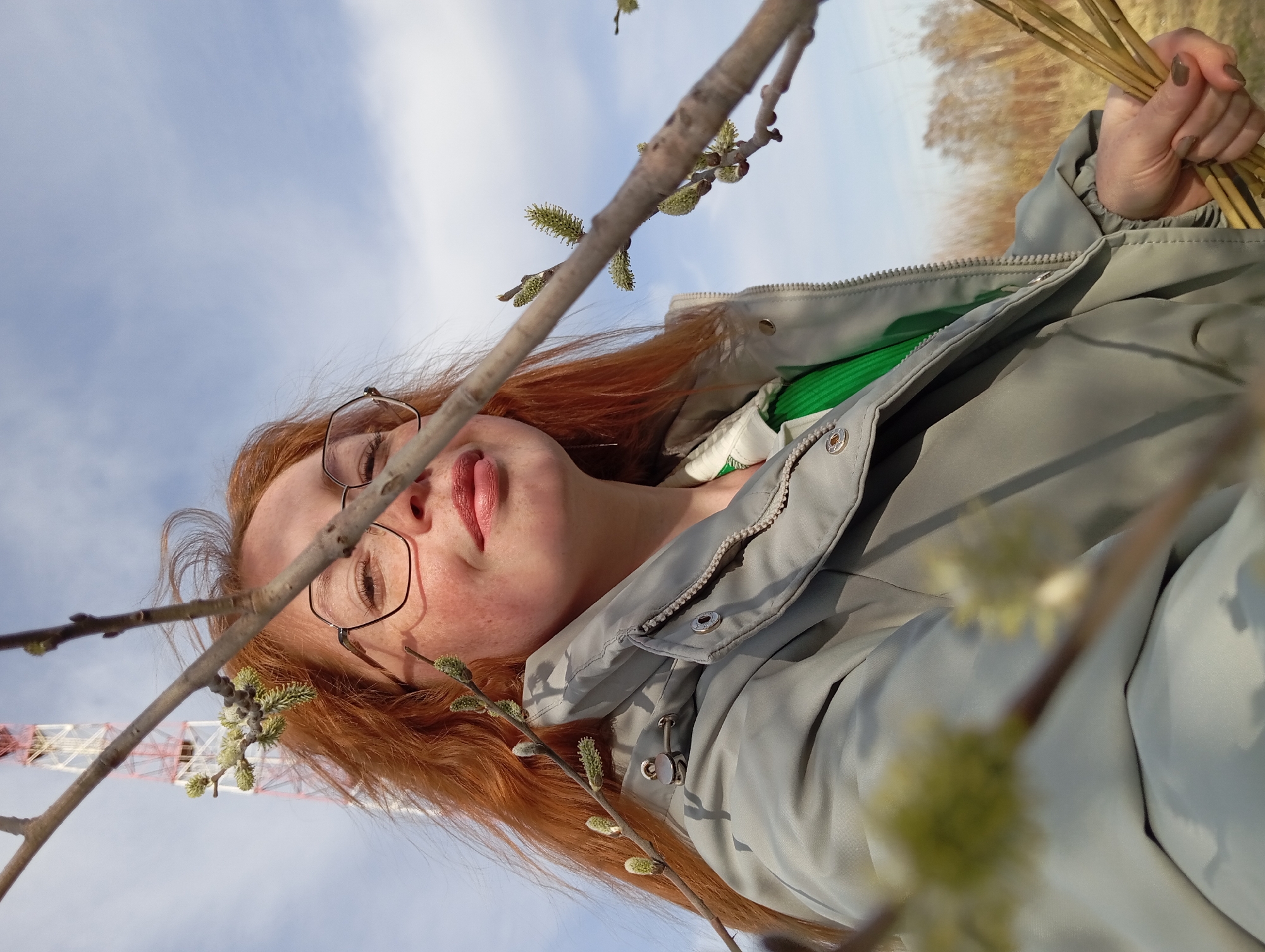 A low-angle outdoor shot shows a red-haired woman wearing glasses and a light gray jacket in Yurgamysh, Russia. In her hand, she holds thin, dry stalks. Emerging branches with budding catkins frame her face. In the blurred background are trees and the pale sky. Behind the branches, a red and white industrial tower is visible.