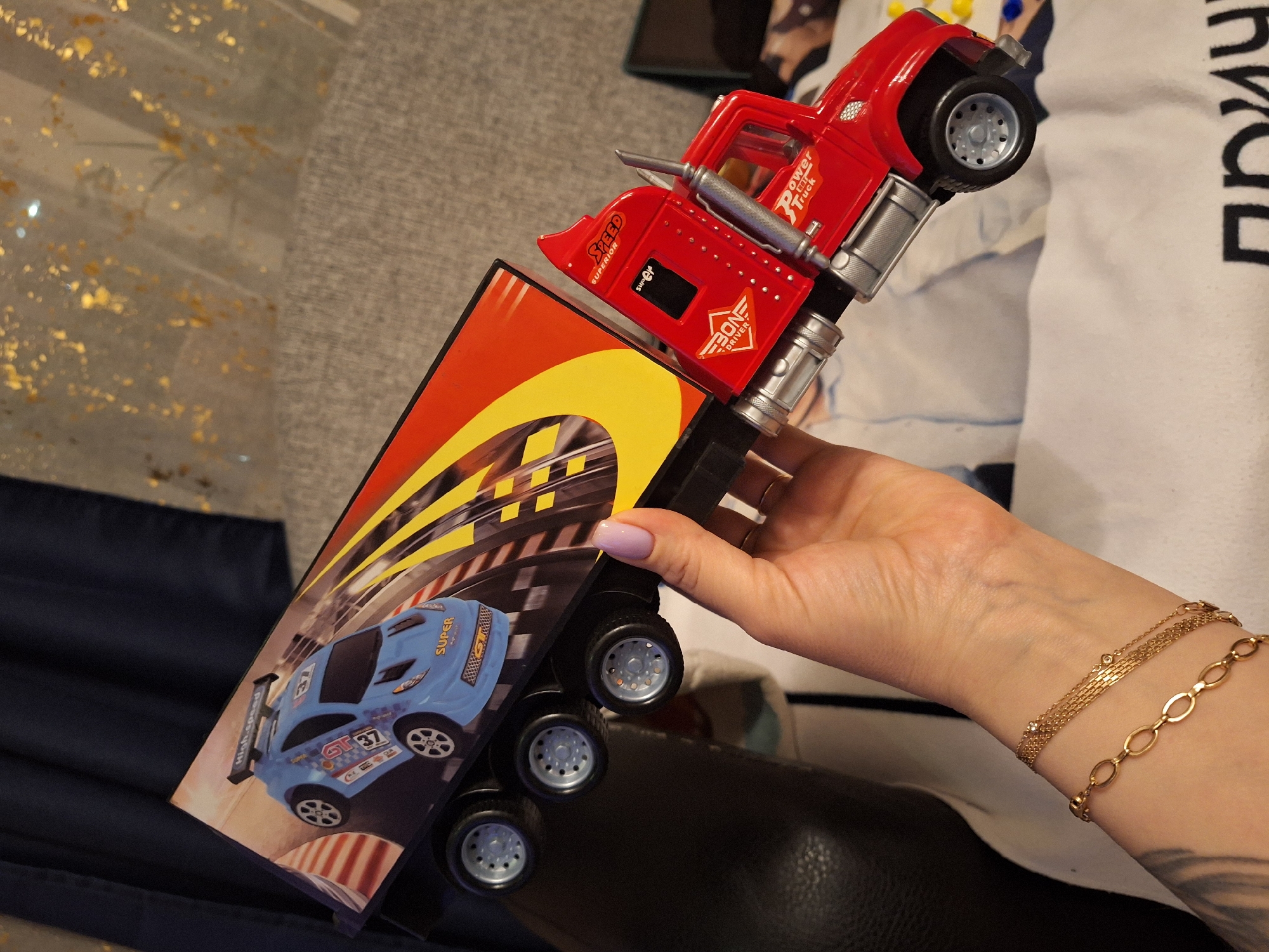 An indoor, close-up shot features a person's right hand holding a red toy semi-trailer truck. The toy is angled upwards, with the truck's cab to the right and a section of its trailer to the left.
The person's hand has fingernails painted with light purple polish. Their wrist is adorned with two gold-colored bracelets: one a delicate chain featuring small, clear stones, and another with larger, interconnected oval links. A dark tattoo is partially visible on the inner forearm below the bracelets.
The toy truck's red cab has silver-colored accents, including exhaust pipes and what appear to be fuel tanks. Text visible on the cab includes "Power Truck," "Speed SUPERIOR," and "BONDRIVER," along with a small black label that reads "SURE." The cab has one visible grey wheel with a silver hubcap. The trailer section of the toy displays a vibrant graphic of a blue racing car, labeled "SUPER GT 37," with additional text such as "High-speed" and "V8 engine," set against a checkered race track with yellow stripes. This trailer segment has three visible grey wheels with silver hubcaps.
The background is largely blurred. On the left, there's a grey fabric with golden, shimmering specks, alongside a dark blue fabric. On the right, a white fabric, possibly a t-shirt, shows large black Cyrillic letters, part of which reads "ПРИ." The scene suggests a casual indoor setting, potentially in Moscow, Russia, as provided by the context. No specific time of day or emotional states are discernible.
