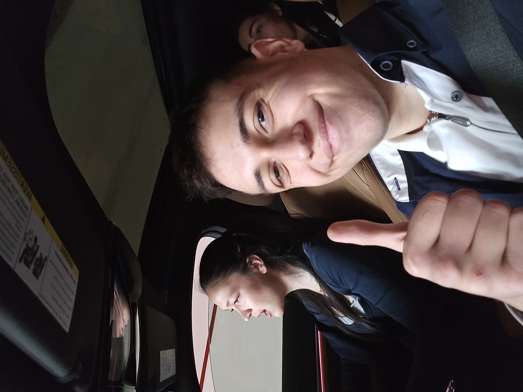 A photo shows two people sitting in a car. One person in the front is smiling at the camera and giving a thumbs-up. Another person is in the back. The location is unknown.