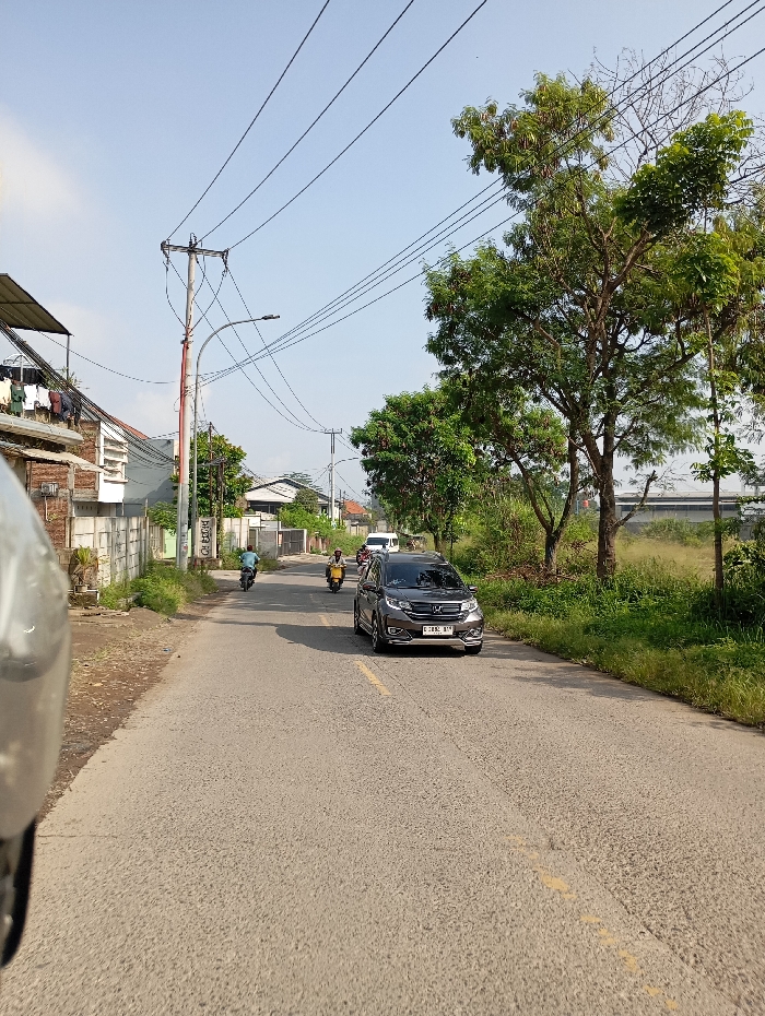 The image depicts a daytime street scene in Pataruman, Indonesia, under a clear blue sky. A two-lane asphalt road, marked with a faded yellow dashed center line, extends into the background.
On the left side of the road, a concrete wall borders residential buildings, some with visible laundry hanging from upper levels. Multiple utility poles supporting numerous overhead power lines and a street light line this side of the road.
The right side of the road features a dirt shoulder transitioning into dense green foliage and several large trees. Beyond this vegetation, an unkempt, open field is visible.
Vehicles are present on the road. In the mid-ground, a dark grey Honda BR-V, bearing Indonesian license plate E 1692 BJ, is positioned slightly right of the center line, facing the viewer. Behind the BR-V, two motorcycles are visible in the left lane; one rider is wearing a yellow jacket. A white vehicle is partially discernible in the far distance.
The perspective is from a low vantage point, with a portion of a grey object, consistent with a motorcycle helmet or vehicle part, visible in the bottom-left foreground. No direct interactions between individuals are discernible, only the movement of vehicles on the road.
