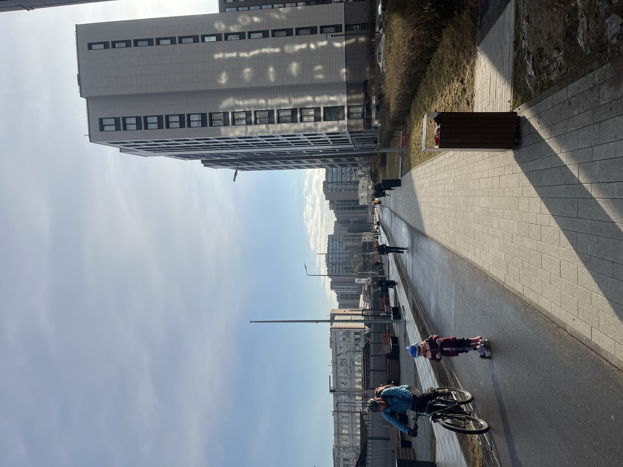 The image shows an urban scene in Saint Petersburg, Russia. In the foreground, a person on a bicycle and a child on roller skates are present. The cyclist is wearing a blue jacket and a helmet, and the child is wearing a helmet and roller skates. They appear to be on a paved path or sidewalk. In the background, there are modern residential buildings. The path is lined with trees and bushes. Other people are walking in the distance. The sky is blue with some clouds. The photograph is taken in the daytime with sunlight casting shadows.