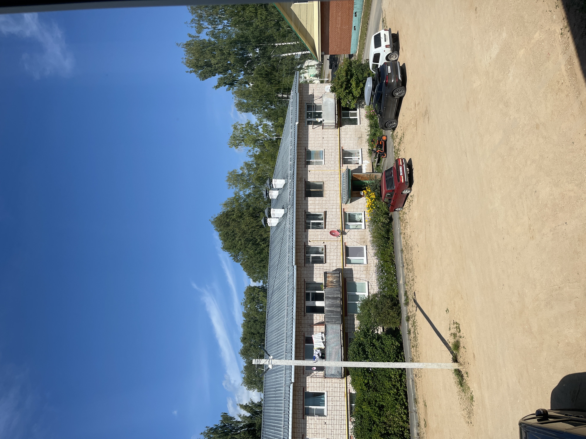 The image shows a multi-story brick building with a corrugated metal roof, likely an apartment building, located in the city of Tarnogsky Gorodok, Russia. In front of the building, there is a dirt parking area with three cars: a red sedan, a dark grey SUV with a roof rack, and a white SUV. A small orange scooter is parked near the entrance of the building. A utility pole stands in front of the building, casting a long shadow. Laundry is hanging on a balcony of the building. Green bushes and trees are visible around the building and in the background. The sky is clear blue with some wispy white clouds. The scene appears to be a residential area on a sunny day.