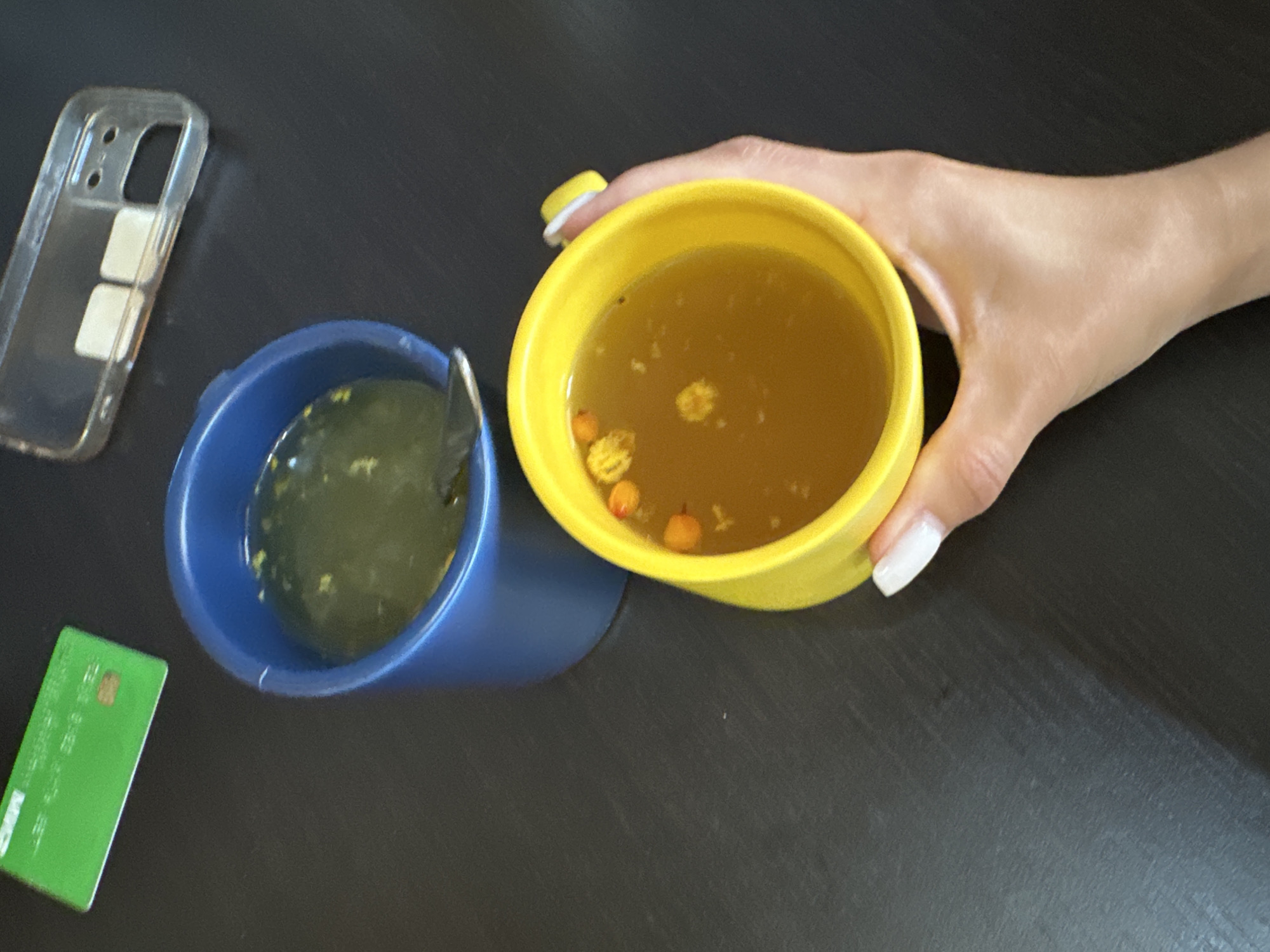 The image depicts a dark, textured tabletop with several objects arranged on it. In the foreground, a person's hand with neatly manicured white nails is holding a bright yellow mug filled with a golden-colored liquid containing small, orange and yellow particles. To the left and slightly behind the yellow mug, a blue mug is visible, also containing a similar liquid with scattered particles. A silver spoon is partially submerged in the blue mug. In the upper left corner, a green card with white text and a small brown chip is partially visible. Further back, a clear plastic phone case with two square, light-colored objects inside is present. The scene is illuminated by overhead lighting, casting soft shadows. The overall setting suggests a casual indoor environment, possibly a home or office.