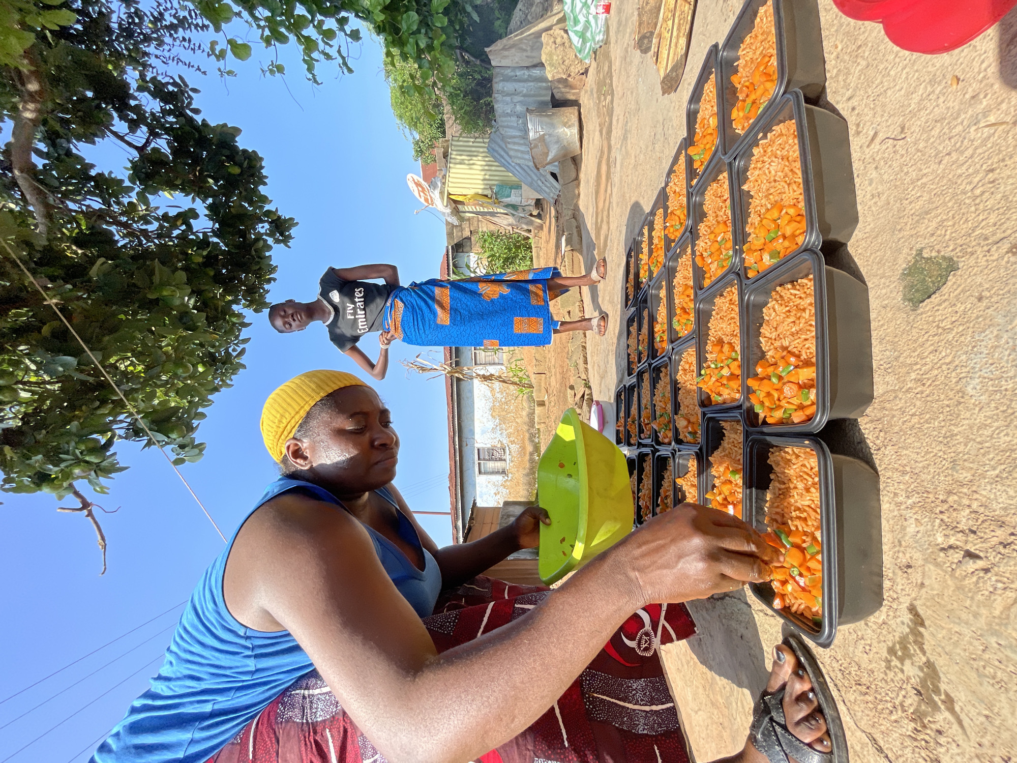 In Jos, Nigeria, a woman in a blue shirt and a red patterned skirt with a yellow head covering is distributing food from a bright green bowl into black rectangular containers. She is hunched over, carefully adding food to one of the containers. In the background, a young woman in a black t-shirt with "Fly Emirates" printed on it and a blue patterned wrapper stands looking towards the camera. The containers, filled with rice and diced vegetables, are arranged in rows on a dirt or concrete surface. The scene appears to be outdoors, with a tree providing shade overhead and buildings in the background. The sky is clear and blue.