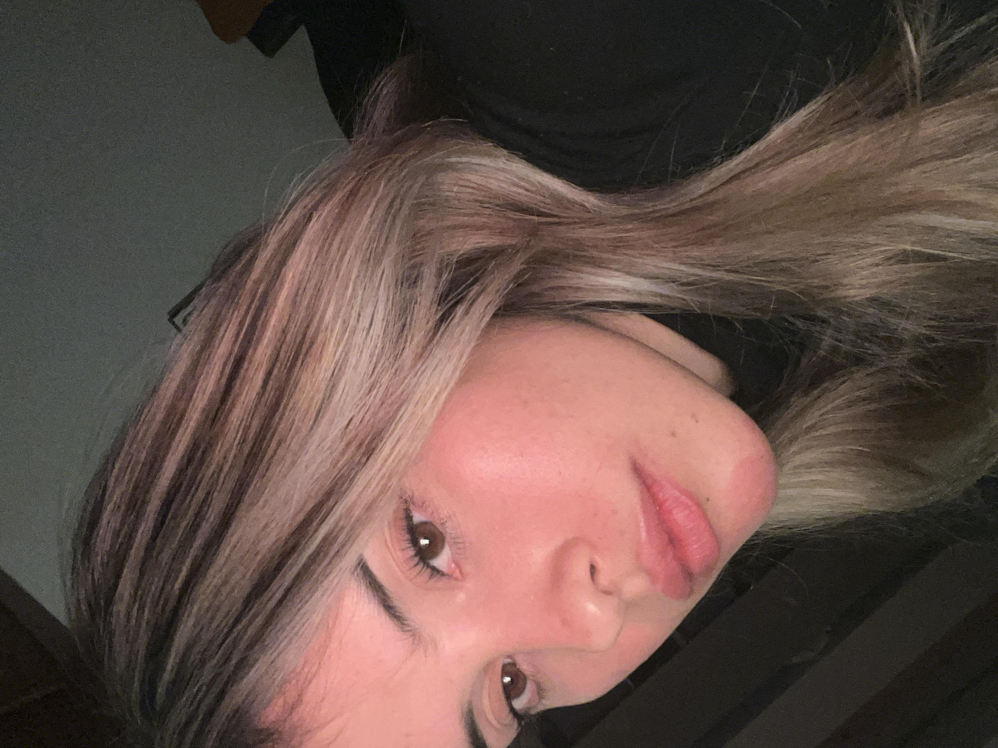 The image is a close-up, low-angle selfie of a young woman with blonde and brown hair, wearing a black turtleneck. She has brown eyes and light skin with some freckles and a reddish tint on her cheeks. Her lips are slightly parted, and she appears to be looking directly at the camera. The background is dark and out of focus, with some hints of brown and dark gray textures. There is no visible indication of the city of Sumalao, Argentina, or any specific urban environment. The overall lighting suggests an indoor setting, possibly at night or in a dimly lit room, with the light source coming from the front and slightly above the subject.
