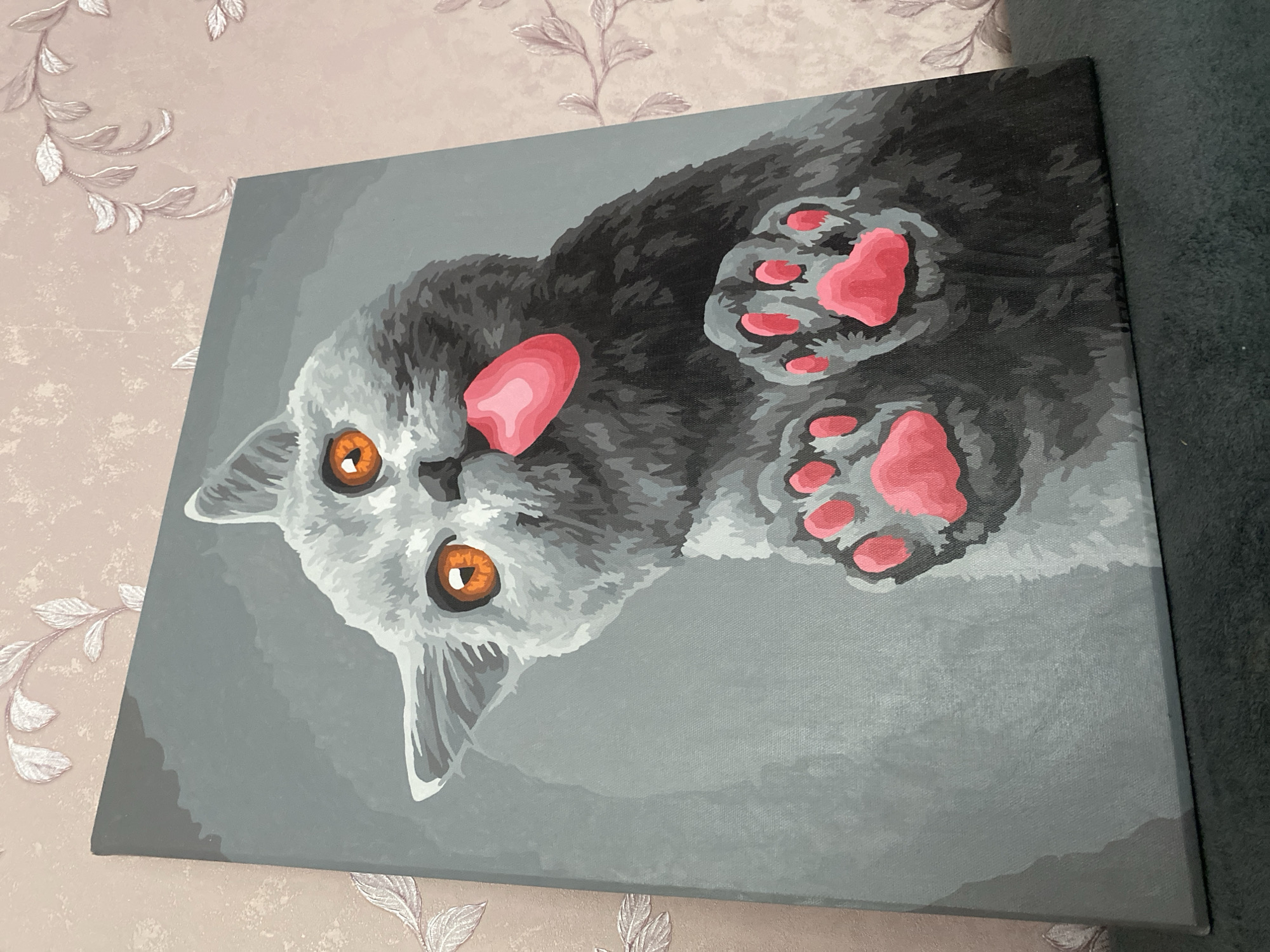 The media file displays a canvas painting of a cat, oriented diagonally. The cat in the painting is depicted lying on its back, looking upwards, with all four paws playfully extended towards the viewer. It features bright, round orange eyes, a soft pink nose/mouth area, and distinct pink paw pads. The cat's fur is rendered in various shades of grey and black, utilizing a stylized, block-of-color technique that gives it a somewhat geometric appearance. The background within the painting is a solid, medium grey.
The painting is set indoors, resting against a dark grey, textured fabric surface, which appears to be part of a sofa or bed. Behind the painting, a wall is visible, covered with light pink or beige wallpaper adorned with a subtle, embossed pattern of leaves or floral motifs. The lighting suggests an indoor setting, but no specific time of day or weather is discernible. There is no visible text in the image. The overall impression is a vibrant and playful artistic representation of a feline in a domestic environment.