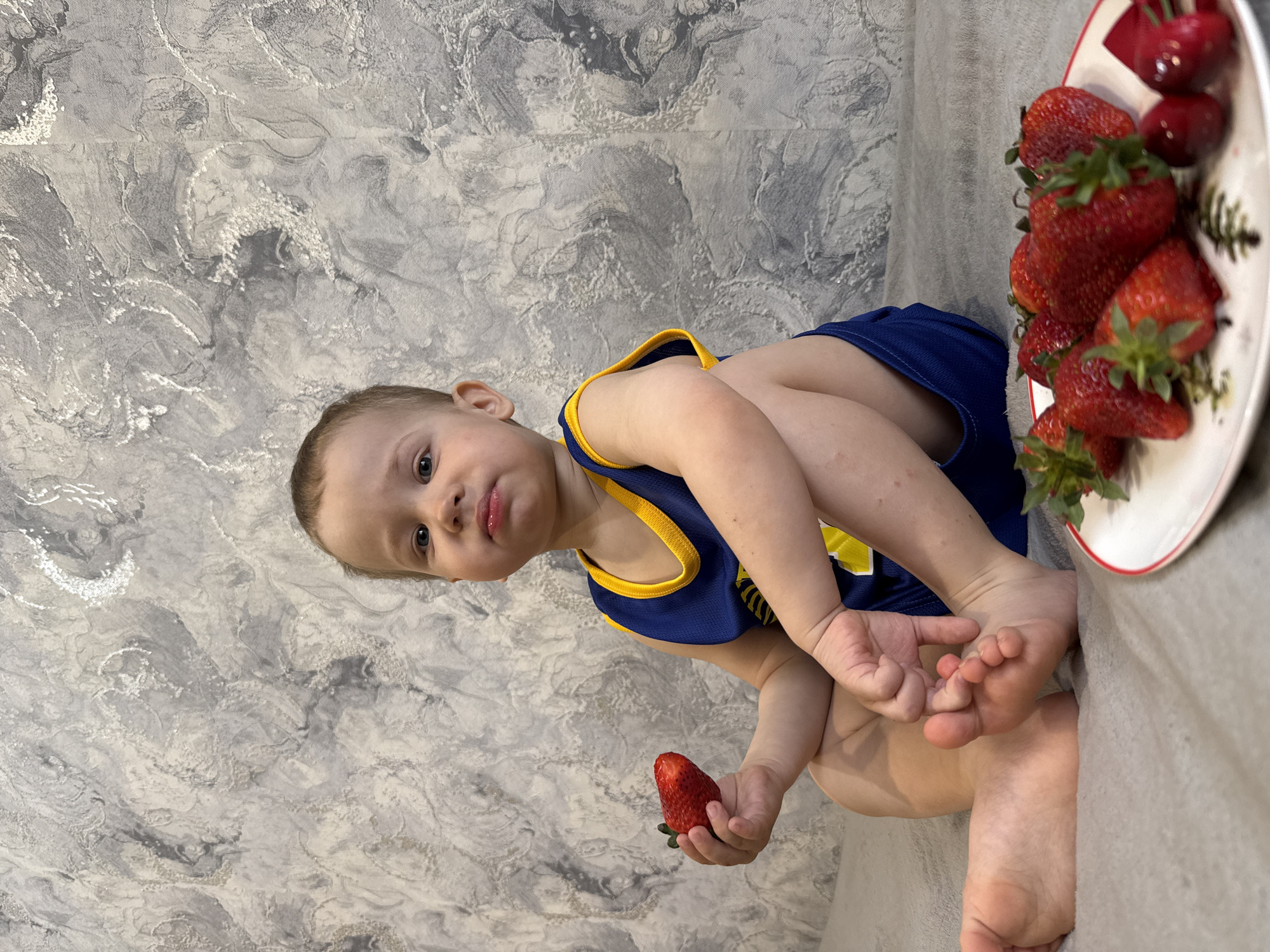 A young child is seated on a light gray surface with a gray and white textured wall behind them. The child is wearing a blue and yellow sleeveless top. They are holding a strawberry in their left hand. A plate of strawberries and cherries is visible to the child's right. The location is Rusanovo, Russia.