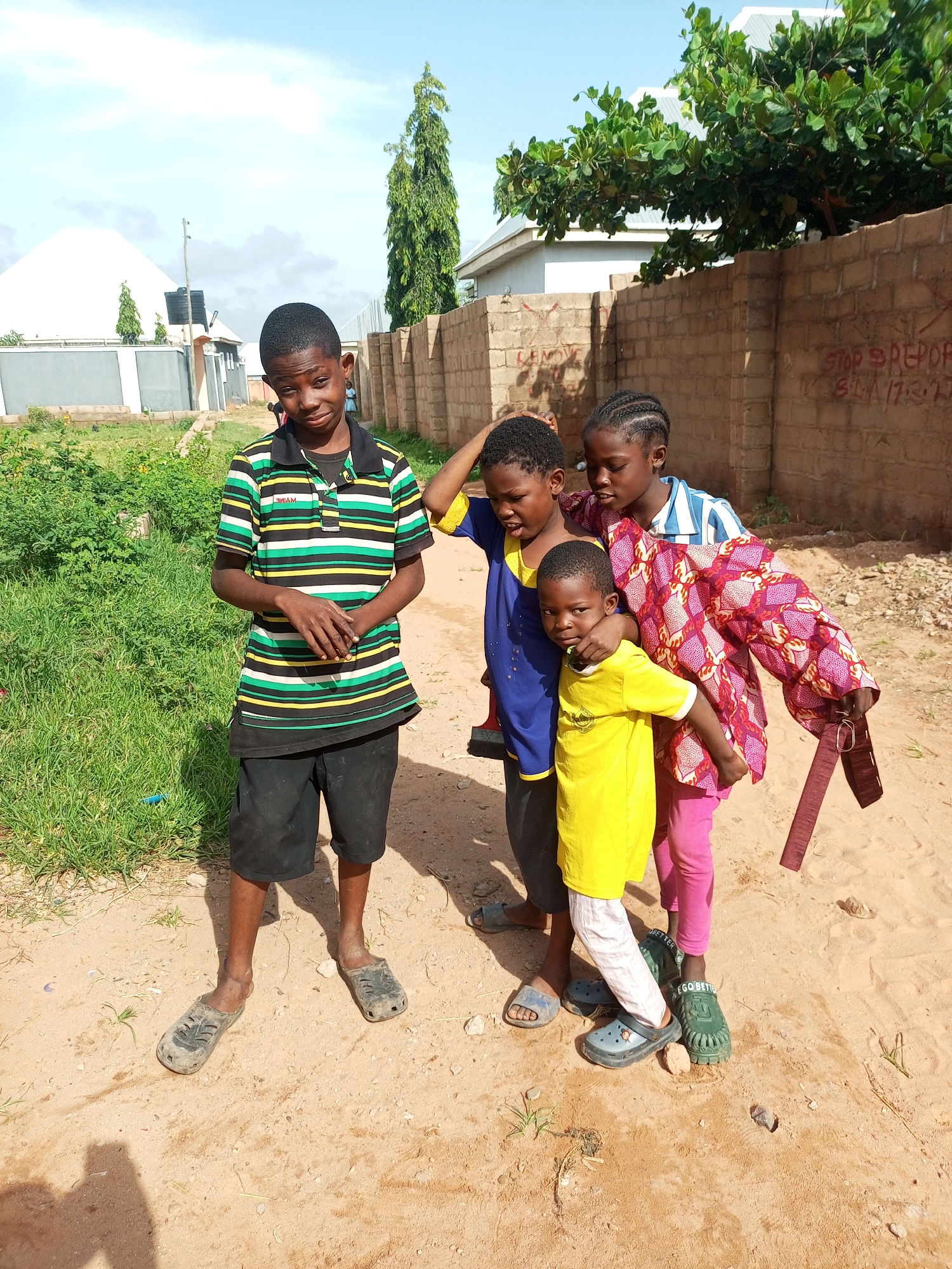 Four children, two boys and two girls, stand on a dirt path in front of a brick wall. The boy on the far left, wearing a striped polo shirt and black shorts, looks towards the camera with a slightly sullen expression. To his right, a second boy in a blue shirt and black pants stands with his arm around the shoulder of a girl. This girl, wearing a patterned red and pink garment over a blue and white striped shirt and pink pants, has her arm around a younger boy in a yellow shirt. The younger boy looks directly at the camera. The background features a brick wall with red graffiti, a tree with green leaves, and buildings. The ground is sandy with scattered dry grass and small stones. The sky is partly cloudy.