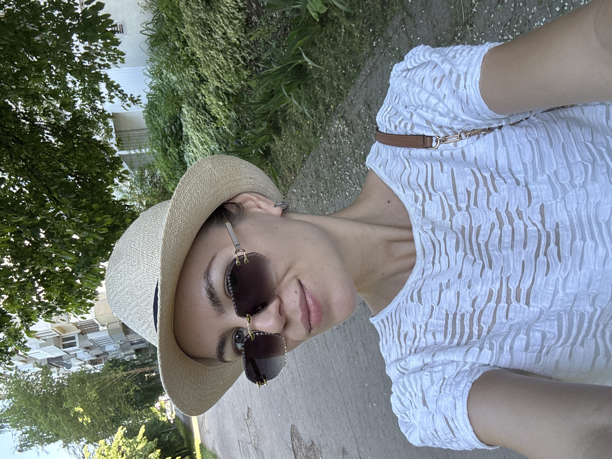 A woman is taking a selfie outside in Unknown, Unknown. She is wearing a white patterned shirt, a straw hat with a navy blue ribbon, and sunglasses with a gold frame. A brown strap hangs over her shoulder. The background shows green trees, grass, and buildings. She stands on a paved walkway.