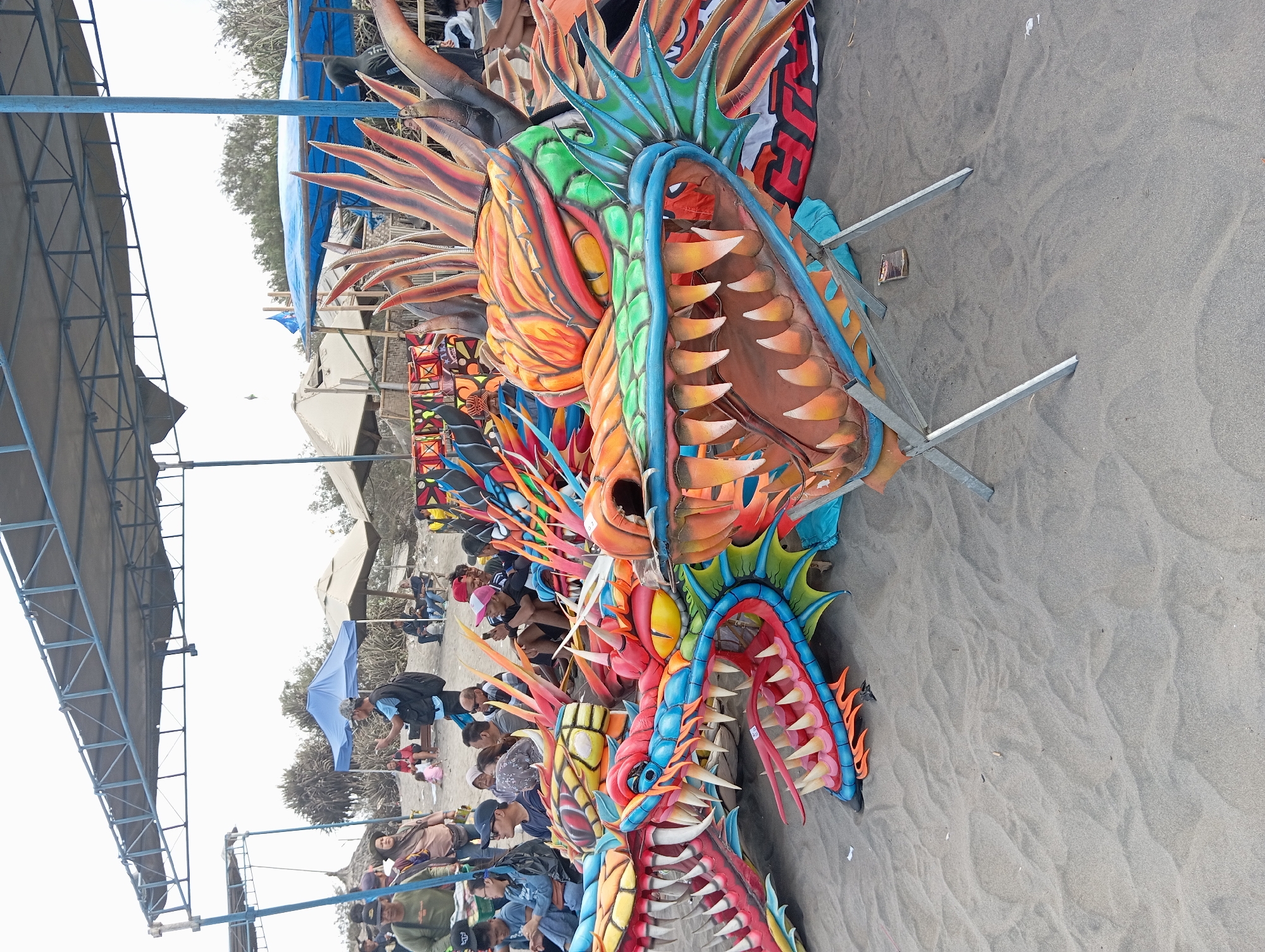 In Parangtritis, Indonesia, on a sandy beach, multiple colorful dragon heads are displayed outdoors. These elaborate props, with their sharp teeth and vibrant scales in shades of orange, red, green, and blue, appear to be part of a festival or performance. Several people are gathered in the background, some sitting and observing, while others stand and interact. Tents and canopies provide shade, suggesting a public event. The overall scene depicts a lively outdoor gathering with a focus on the visually striking dragon decorations.