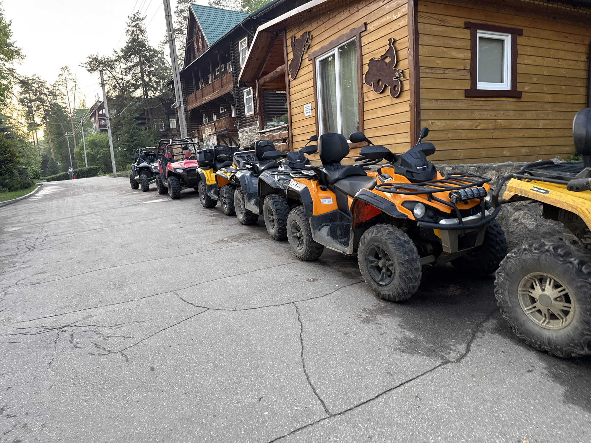 The image depicts a row of ATVs parked on an asphalt road next to a wooden building with a window and a decorative wooden motorcycle cutout on the wall. The location appears to be Zolotoi Plyazh, Russia. The ATVs are of various colors, including orange, yellow, red, and black. The road is cracked, and there are trees and other buildings visible in the background. A person is seen walking on the road in the distance.