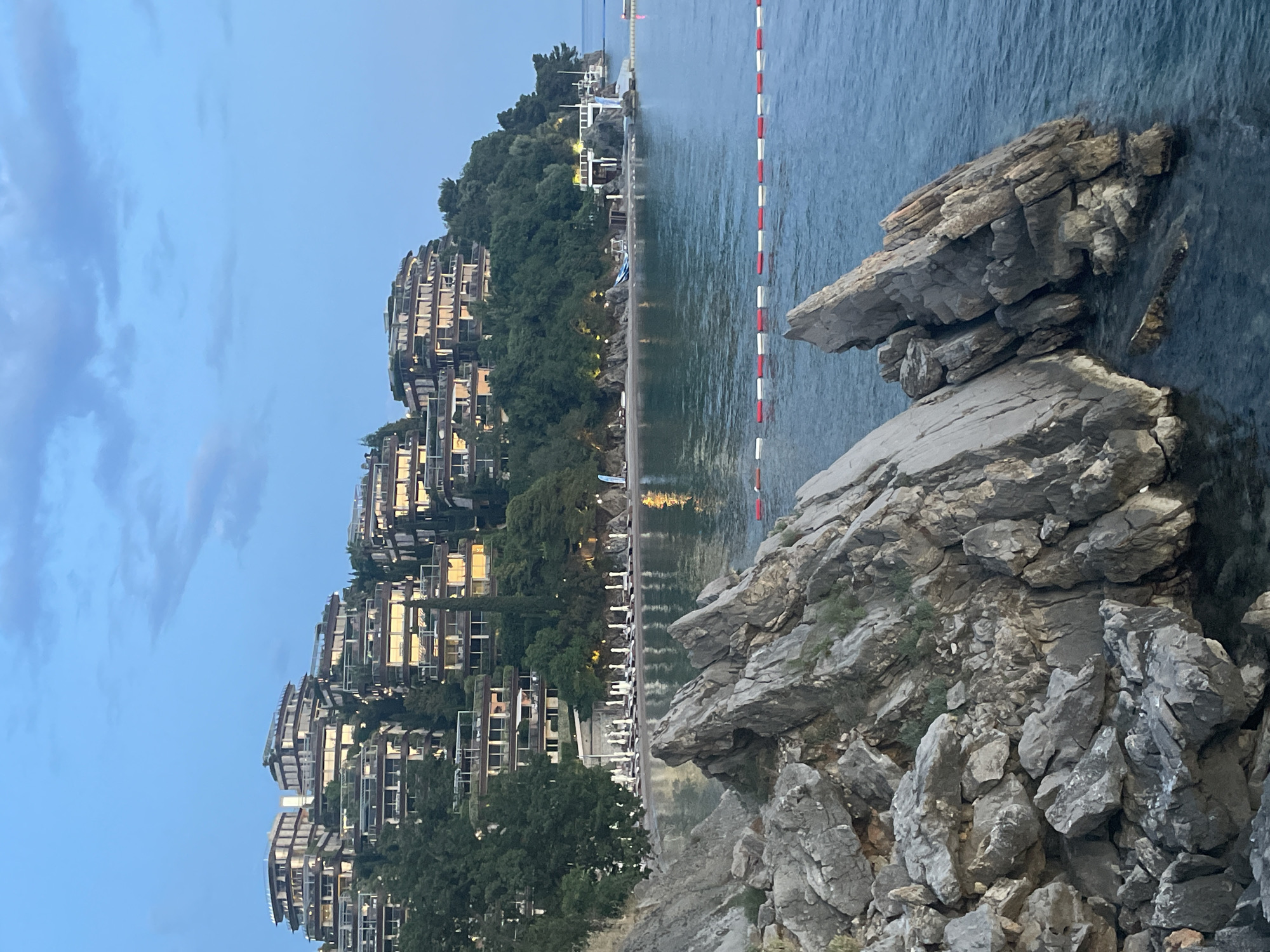 The image depicts a coastal view in Budva, Montenegro. A multi-tiered building complex is built into a hillside covered with trees. The buildings have multiple balconies and appear to be apartments or hotel rooms. In the foreground, there are large rocky formations extending into the water. A beach lines the coast with rows of sun loungers and umbrellas visible. A line of red and white buoys extends into the water. The sky is clear with some clouds, indicating either late afternoon or early morning light.