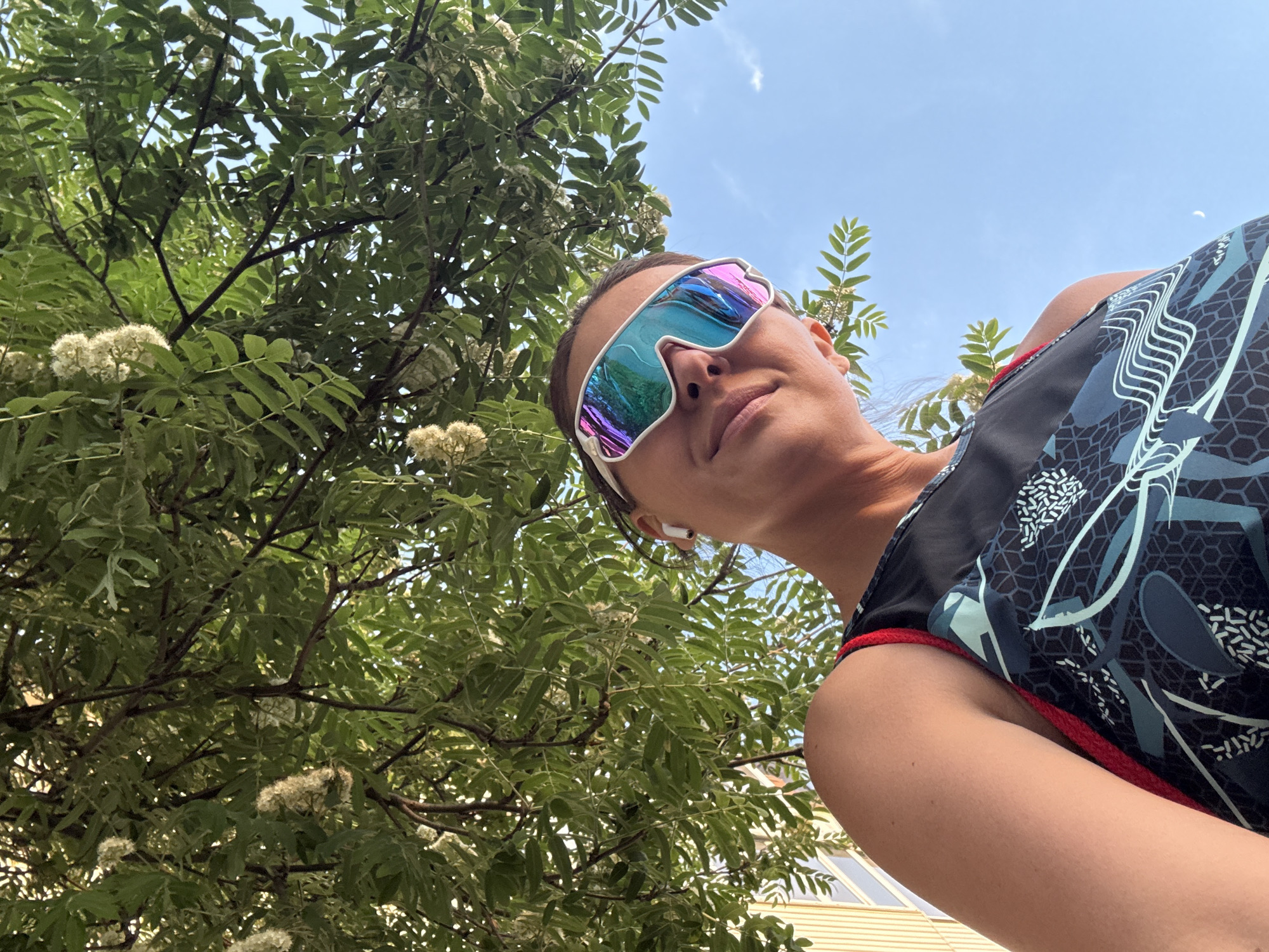 A low-angle shot captures a woman wearing iridescent sunglasses and an athletic top with a geometric pattern. She appears to be near a tree with green leaves and white flowers, partially obscuring a light-colored building in the background. The sky is visible in the upper right corner. This image was taken in the city of Chaikovsky, Russia.