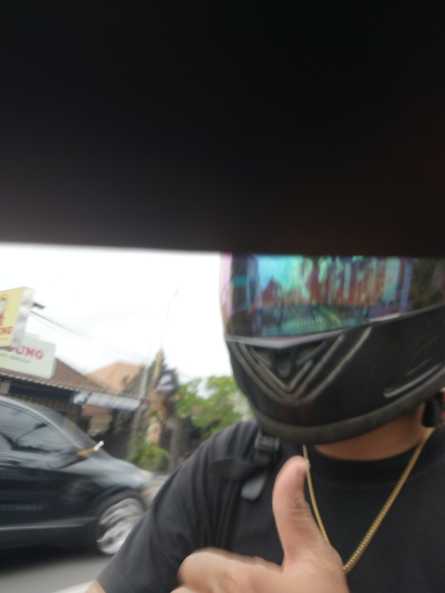 A person wearing a black motorcycle helmet and a black t-shirt with a gold chain is giving a thumbs-up. The helmet has a reflective visor that shows a blurry reflection of buildings and possibly trees. In the background, a black car is visible, along with buildings and signage. One sign in the background is yellow with red lettering that reads "UNG". Another sign is white with red lettering that reads "UNG". The context suggests this image was taken in Denpasar, Indonesia, likely on a street given the presence of the car and buildings.