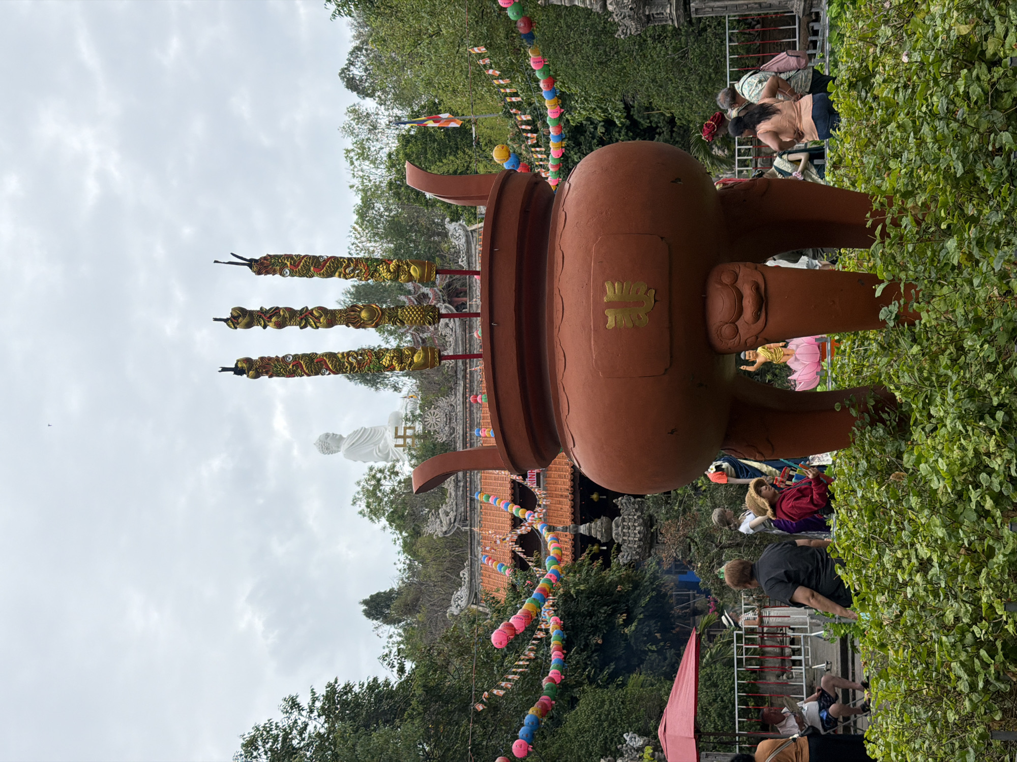 Captured in Tay Nha Trang Ward, Vietnam, the image features a large, reddish-brown three-legged incense burner, the primary subject, dominating the foreground. Sticks of decorative carvings resembling dragons protrude from the burner’s top. A white Buddha statue is visible in the background, positioned on a building with a red-tiled roof. Colorful lantern strings decorate the scene. People populate the midground, some of them appearing to be relaxing or taking photos. Lush greenery surrounds the area. The sky is overcast.