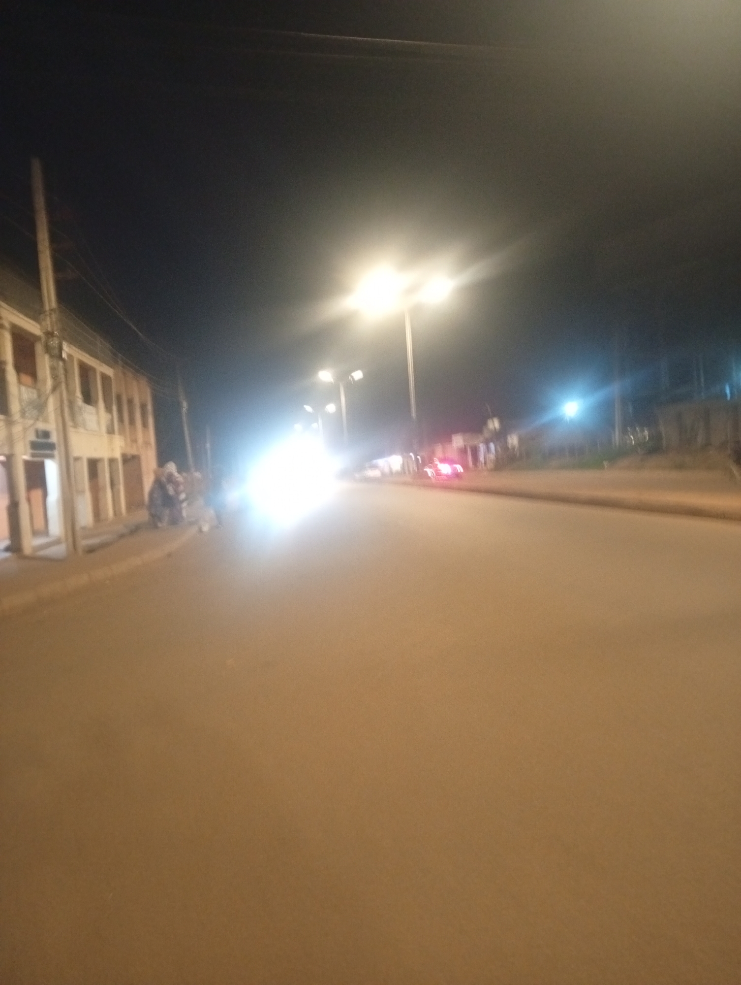 The image shows a street scene at night in Bukuru, Nigeria, illuminated by streetlights and the headlights of a vehicle. A two-story building with multiple windows and a balcony is visible on the left side of the street. In front of the building, several individuals appear to be standing or sitting near a parked motorcycle. The street itself is paved and stretches into the distance, with more streetlights and buildings lining the opposite side. A vehicle with bright headlights is approaching from the right, creating a glare that partially obscures the scene. The overall atmosphere is dark and somewhat blurry due to the low light conditions.